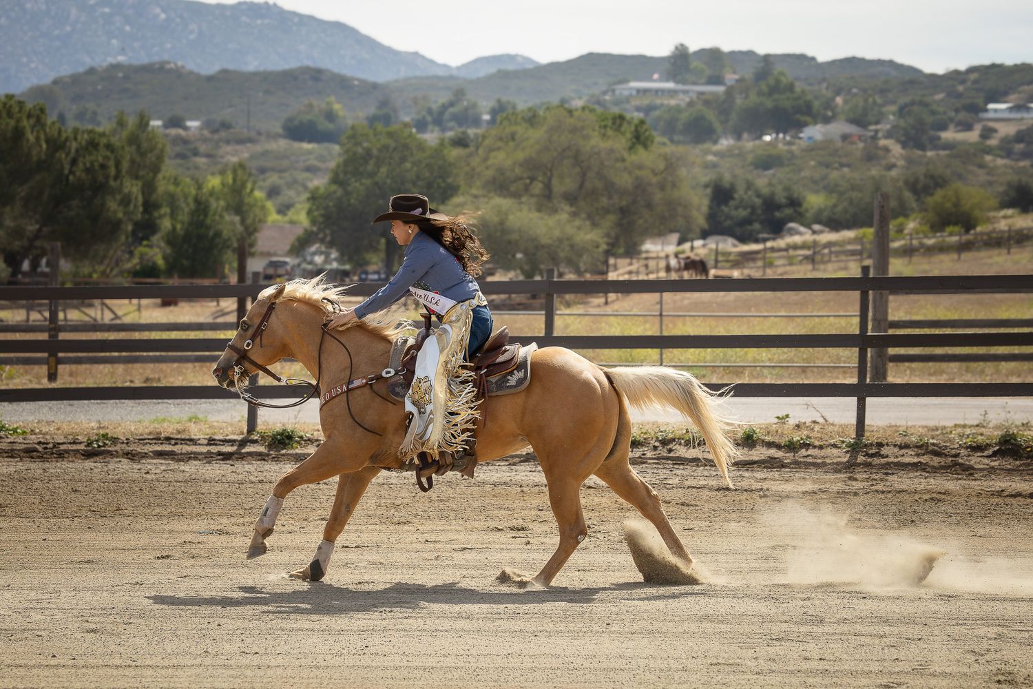 Woman riding a palomino horse in a sandy outdoor arena with mountains and trees in the background.