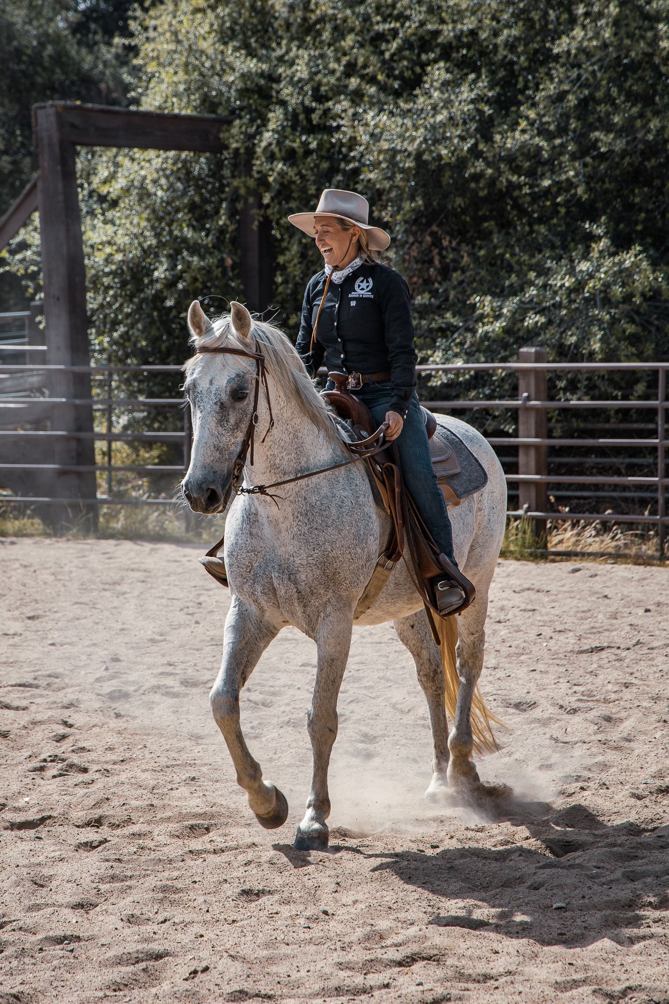 Smiling cowgirl in black shirt and hat riding a white horse in a sandy outdoor arena.