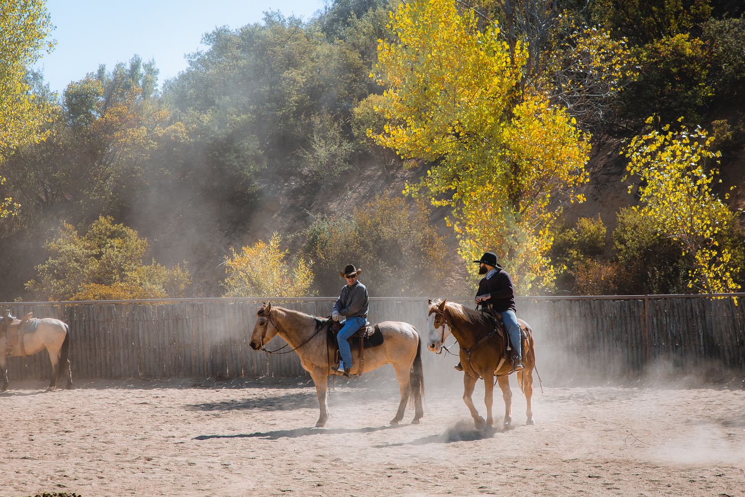Two cowboys on horseback riding through a dusty corral surrounded by autumn foliage.