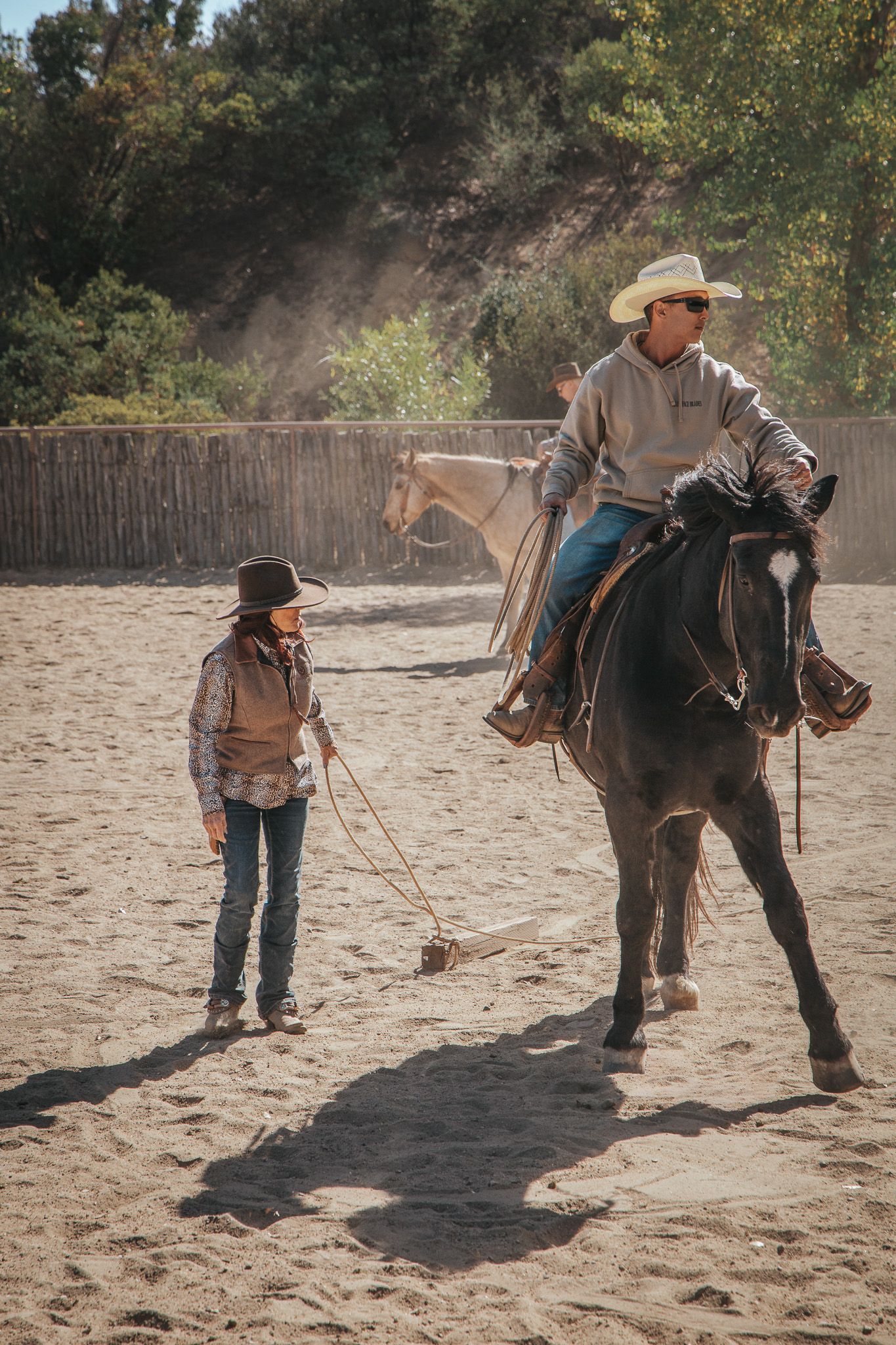 A cowboy on horseback works with a young child learning to rope in a dusty outdoor riding arena.