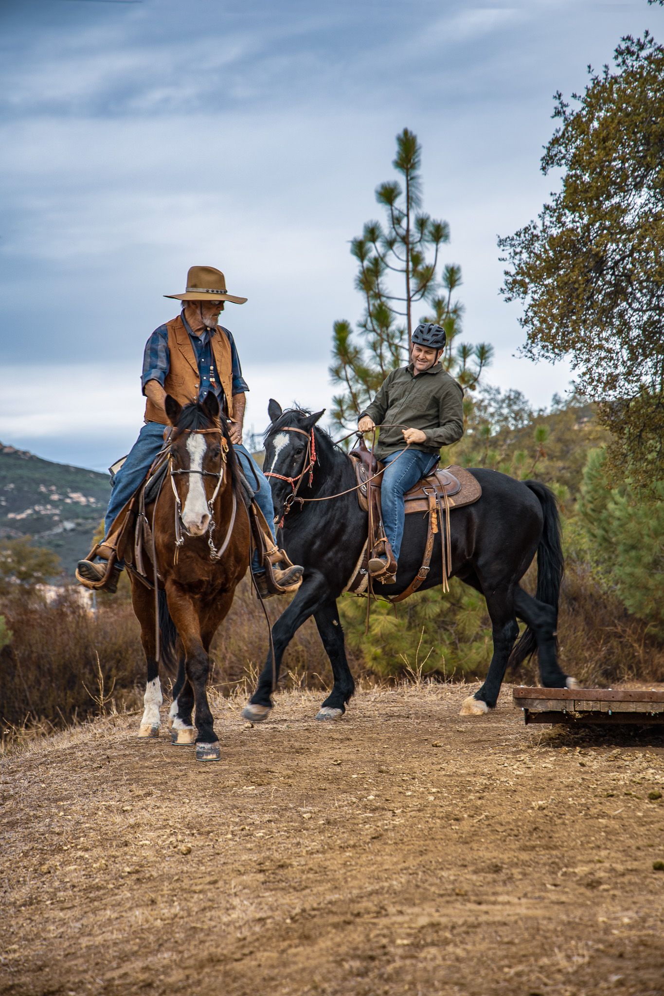 Two riders on horseback, one on a chestnut and one on a black horse, ride together on a scenic trail.
