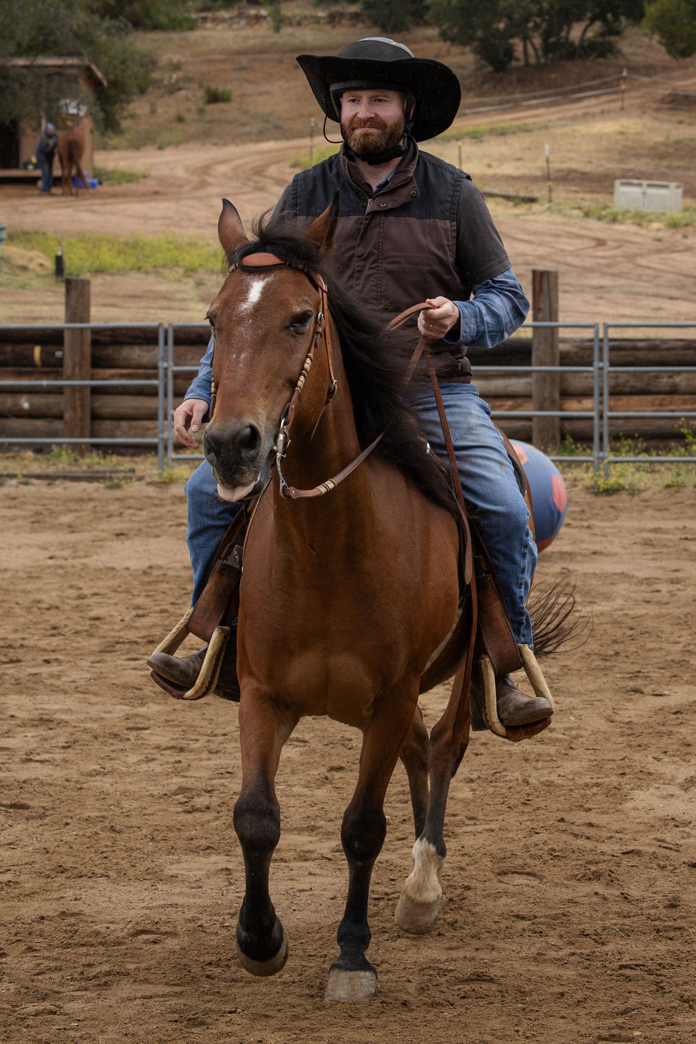 Bearded cowboy in black hat rides brown horse in sandy rodeo arena, smiling confidently during western equestrian event.