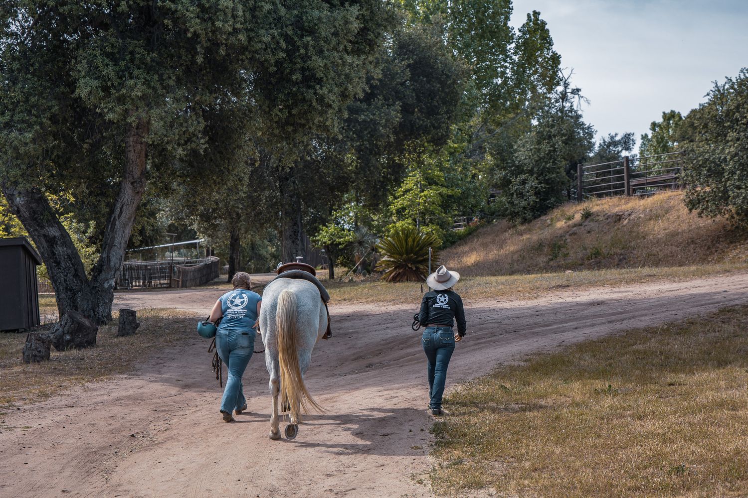 Two people walking a white horse down a dirt path at a ranch surrounded by trees.