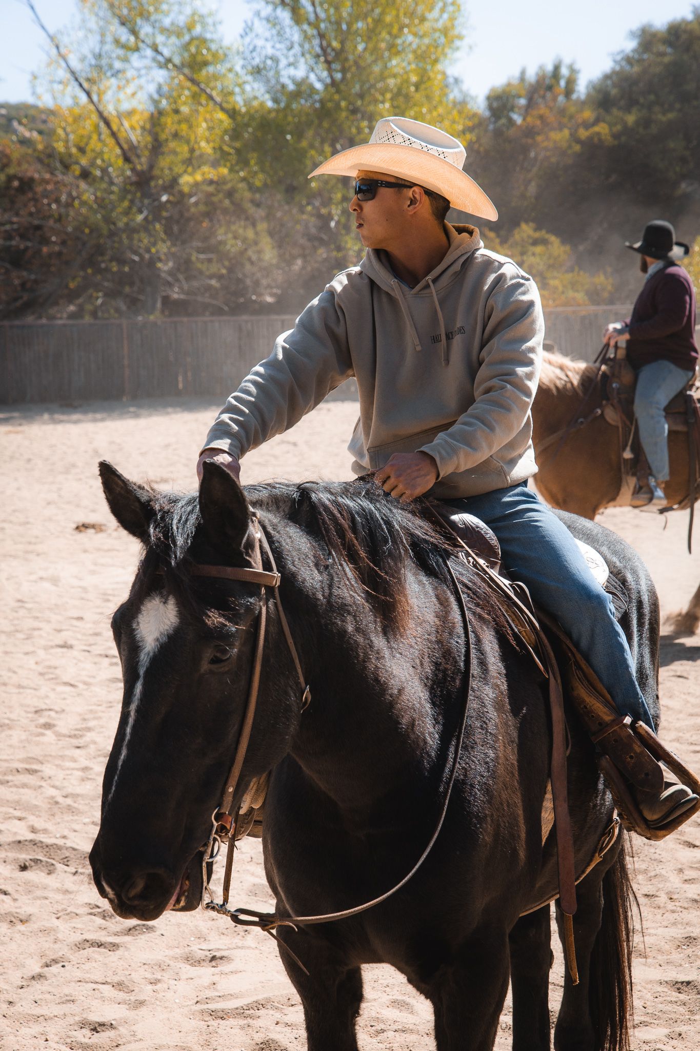 A young man in a cowboy hat and sunglasses rides a black horse in a sandy outdoor riding arena.