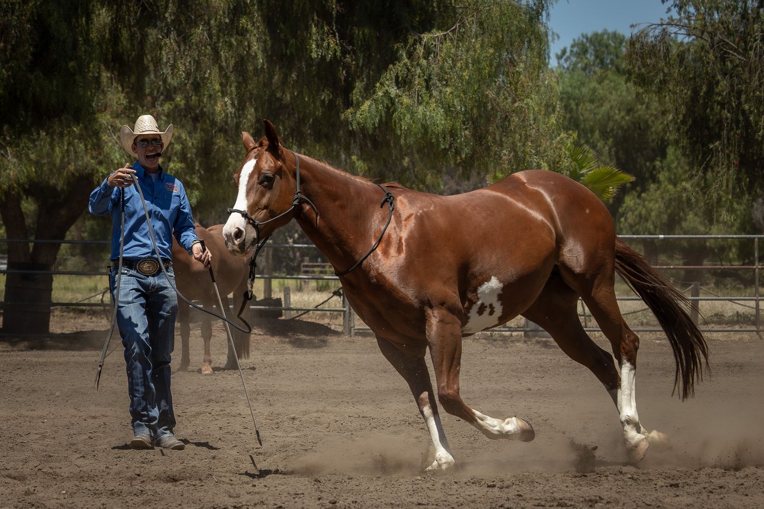 A cowboy in a blue shirt and hat leads a chestnut horse in a sandy arena surrounded by trees.