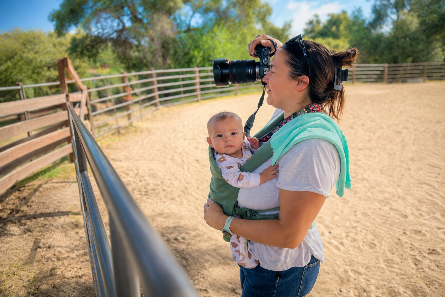 Woman with baby in carrier photographs horses at a sunny outdoor ranch corral with wooden fencing.