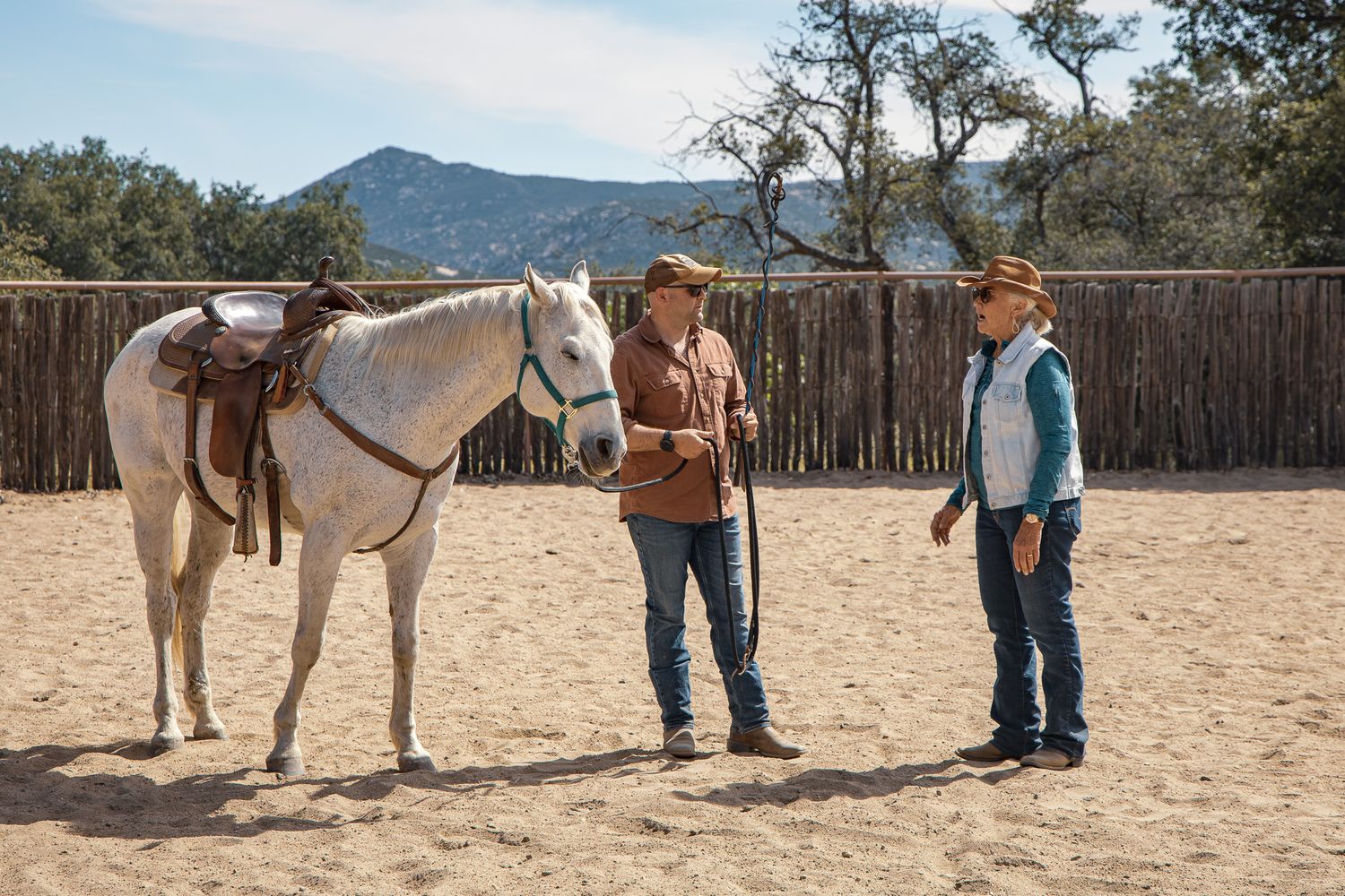 Two cowboys in hats standing with a saddled white horse in a sandy corral with wooden fence.