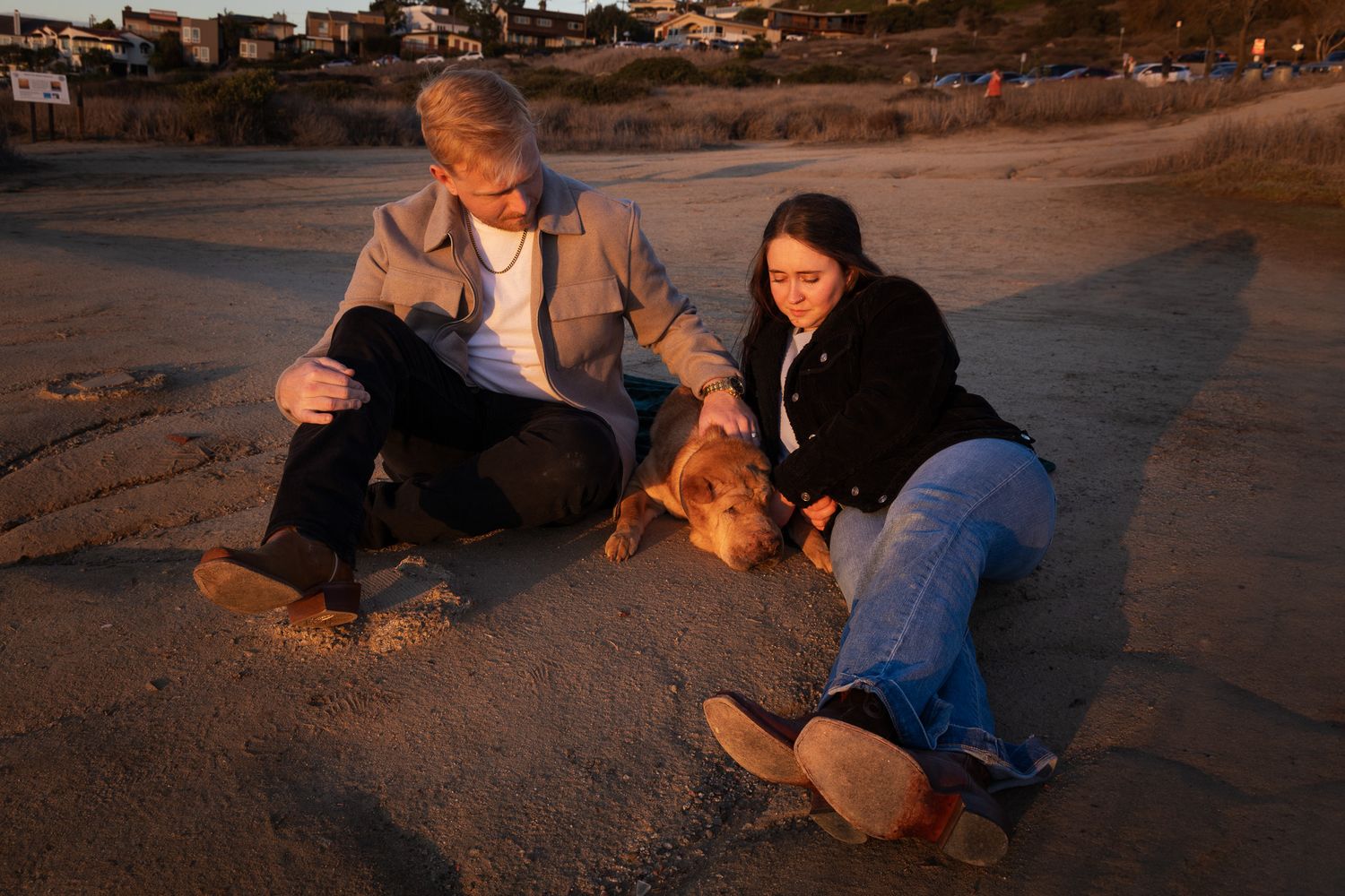 Man and woman sitting on sandy beach petting a brown dog in warm evening light.