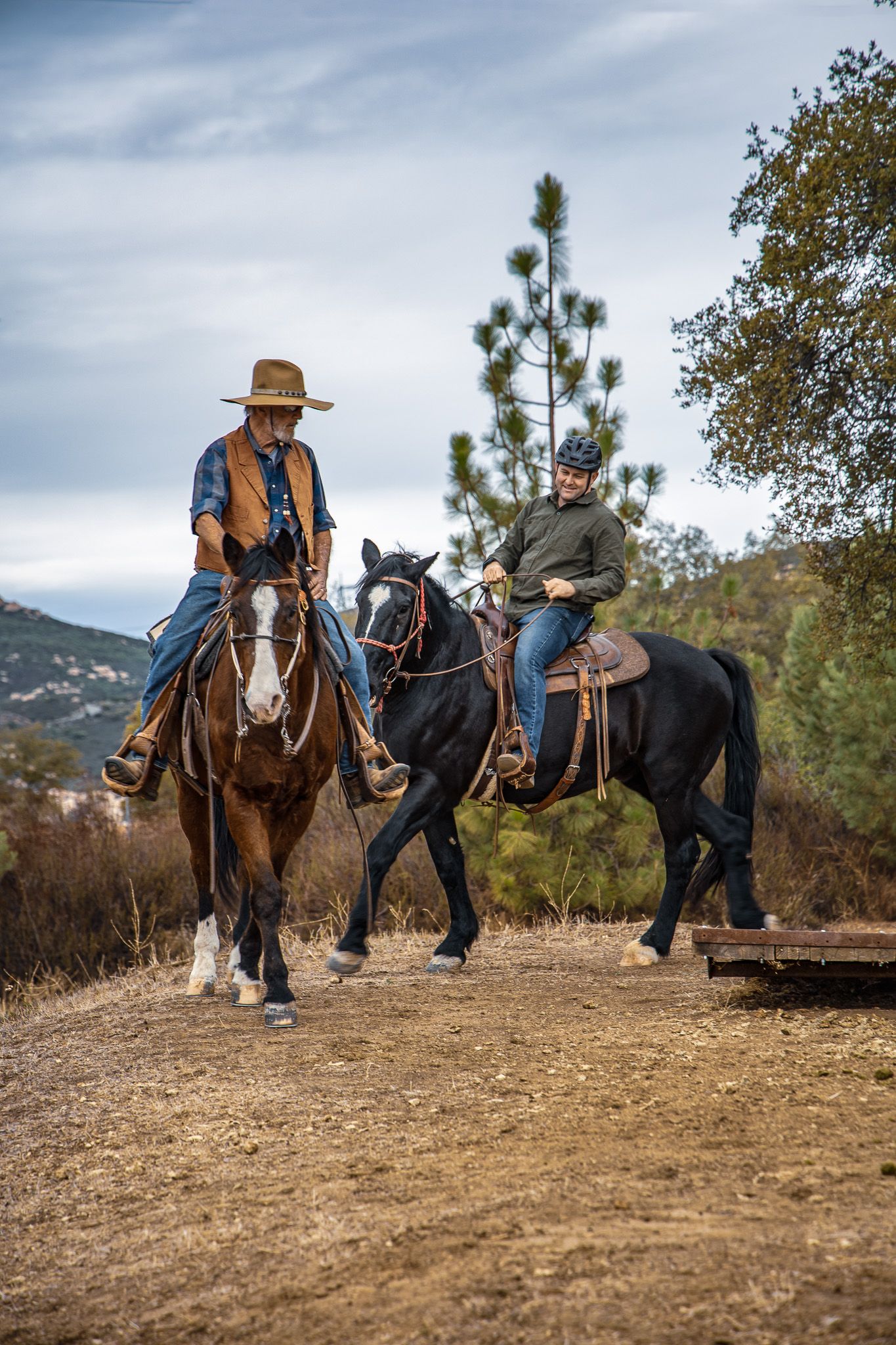 Two riders on horseback, one on a brown and one on a black horse, riding in a rural landscape.