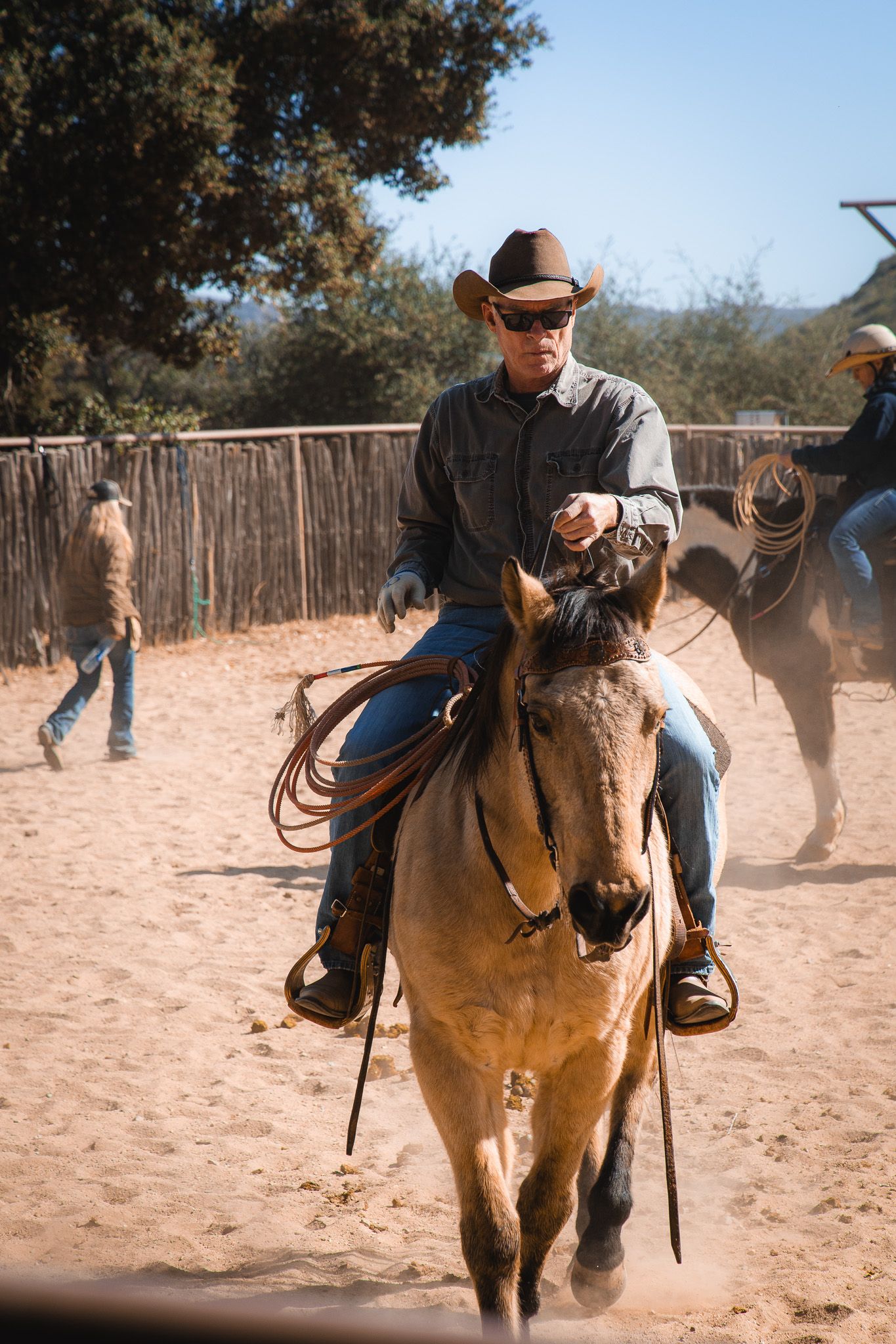 Cowboy in hat and sunglasses riding a horse in a sandy rodeo arena while holding a lasso rope.