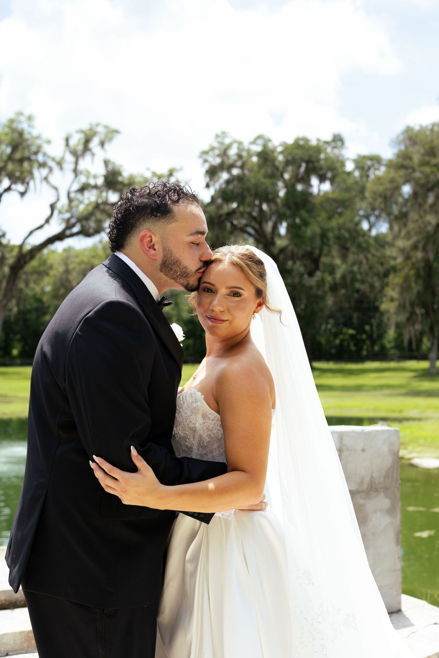 Groom in black tuxedo kissing bride in white gown and veil outdoors near pond with oak trees.