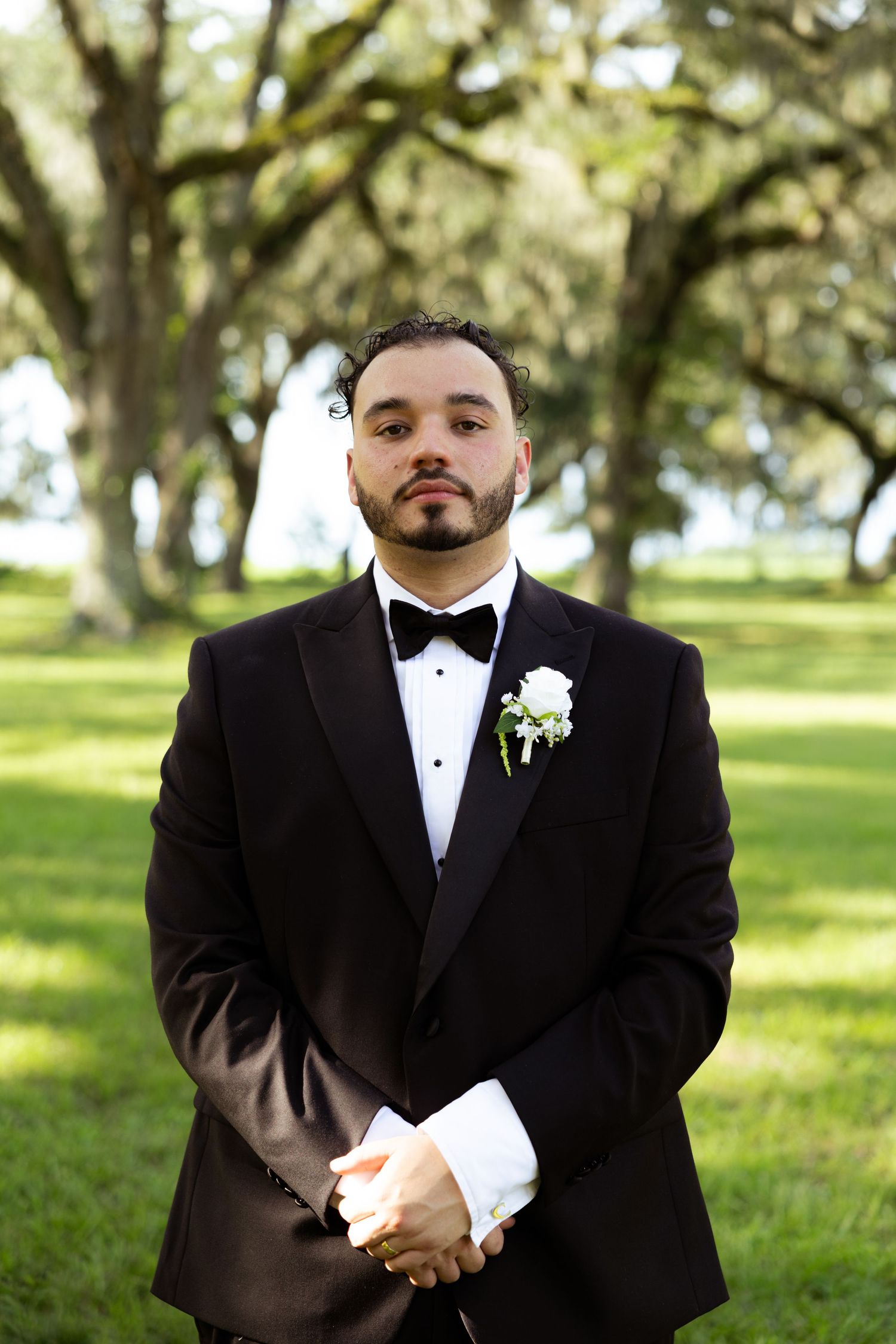Groom in black tuxedo with bow tie and white boutonniere poses outdoors under moss-draped oak trees.