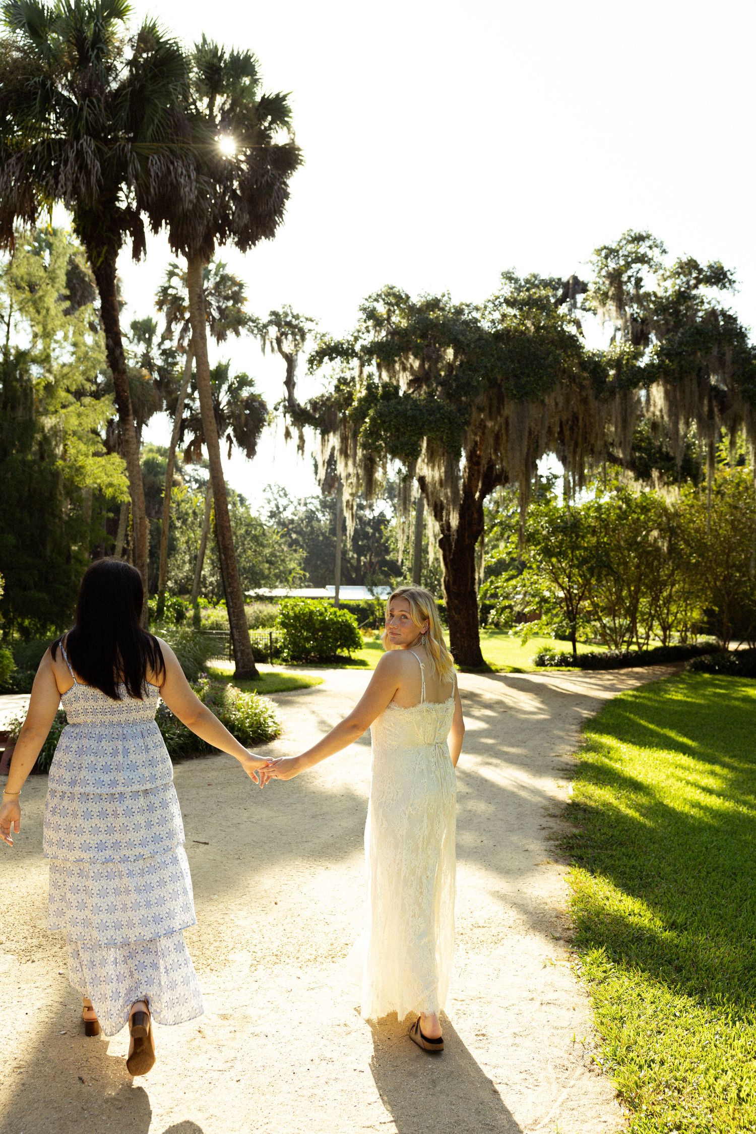 Two women in summer dresses hold hands walking down a sunny park path lined with palm trees.