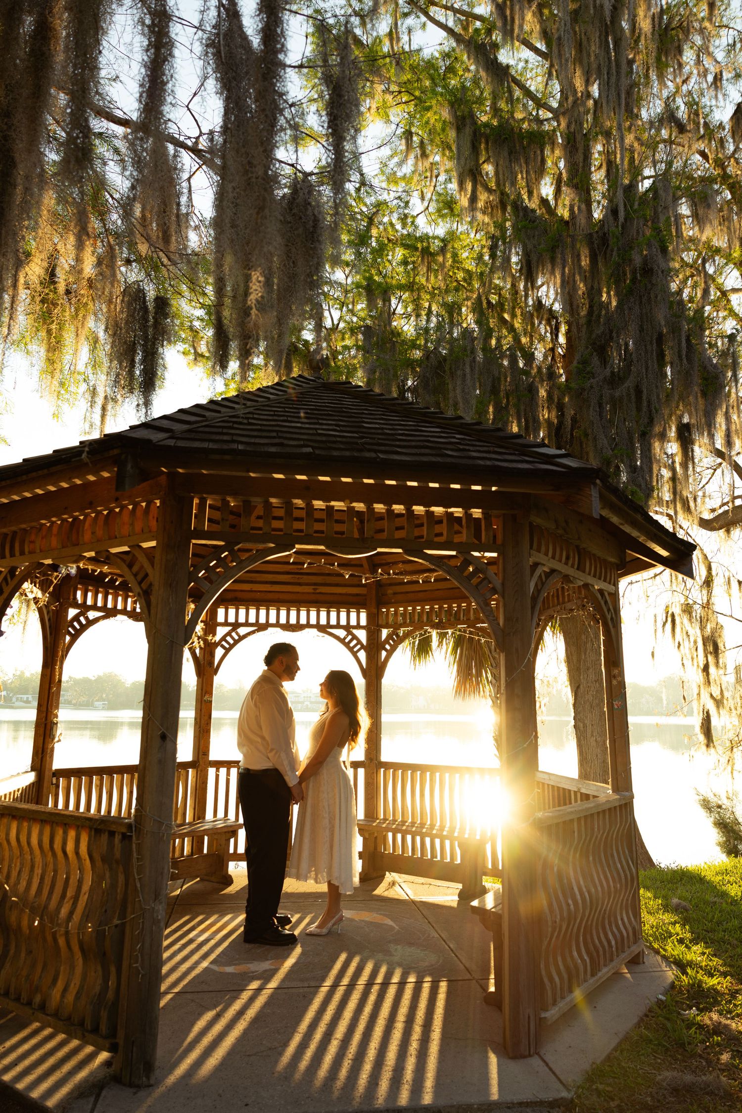 Couple sharing a romantic moment in a wooden gazebo at sunset, with Spanish moss trees and a serene lake backdrop.