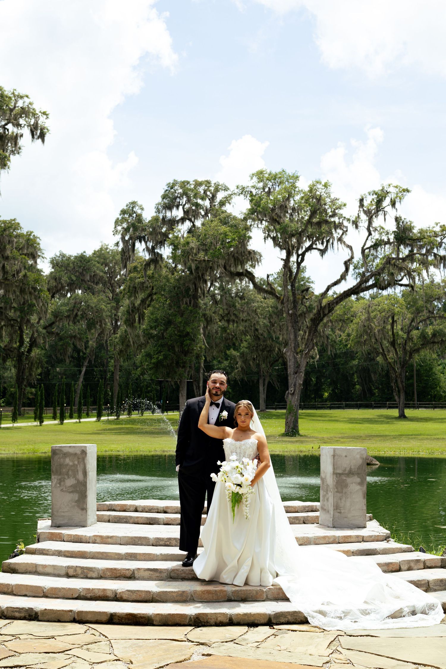 Bride and groom pose on stone steps by a serene pond surrounded by moss-draped trees at an outdoor wedding.