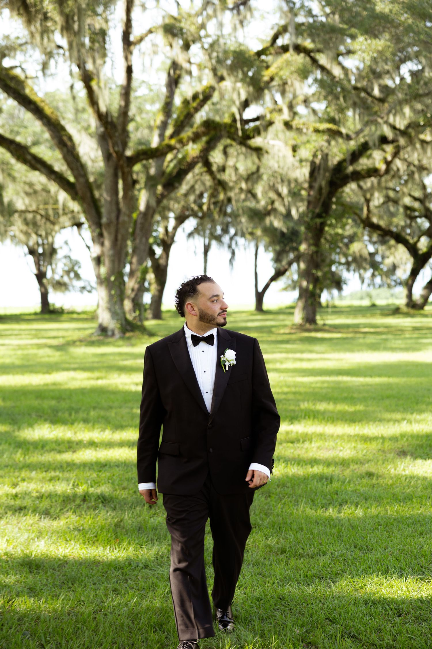 Groom in black tuxedo with white boutonniere walks through lush green lawn beneath sprawling oak trees.