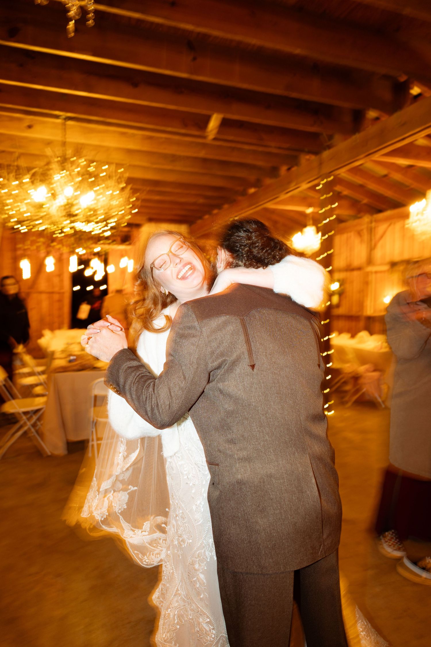Smiling bride in a lace gown and veil dances with her groom in a tweed jacket inside a rustic wooden barn lit by glowing chandeliers.