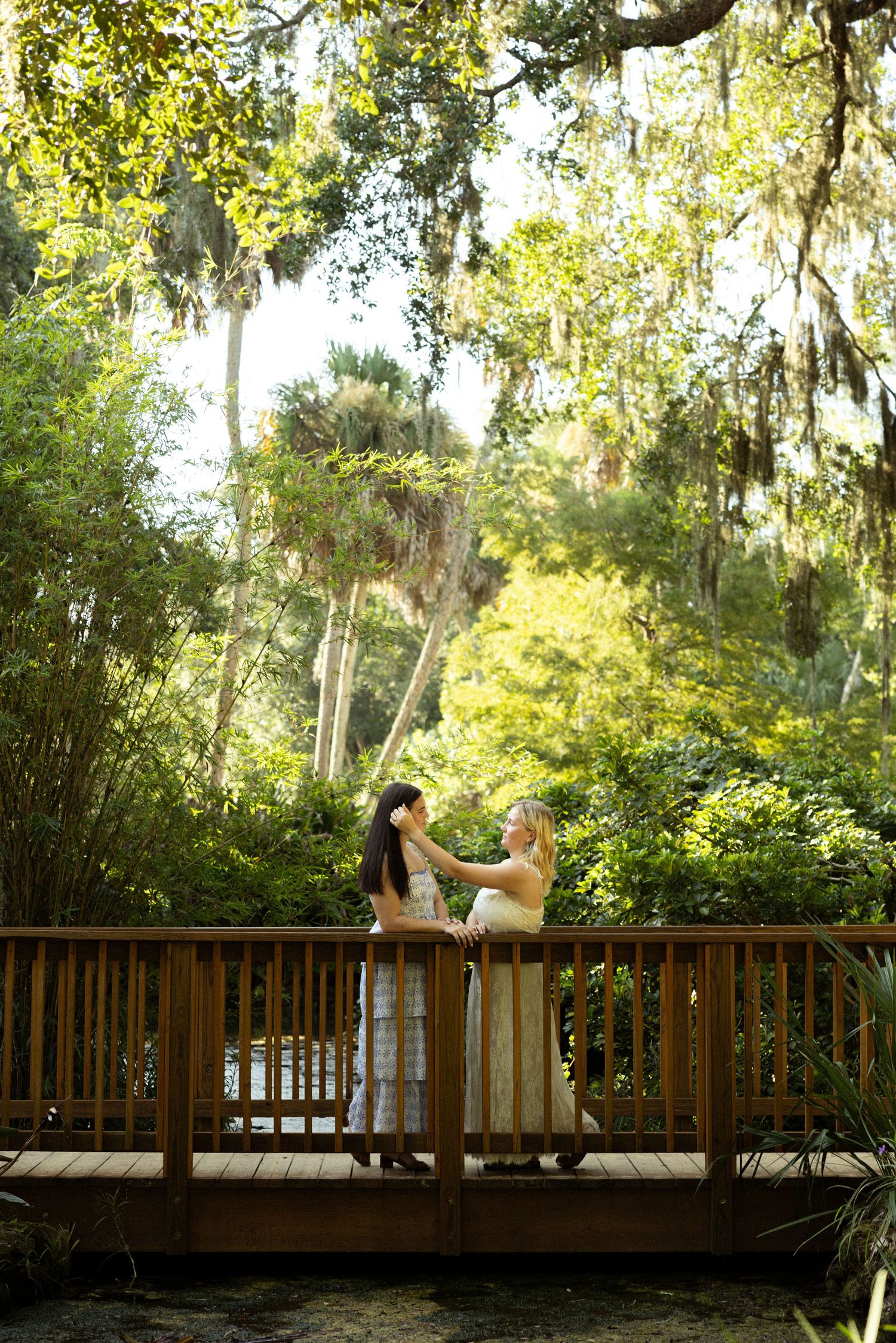 Two women in white dresses share a romantic moment on a wooden bridge surrounded by lush green trees draped with Spanish moss.