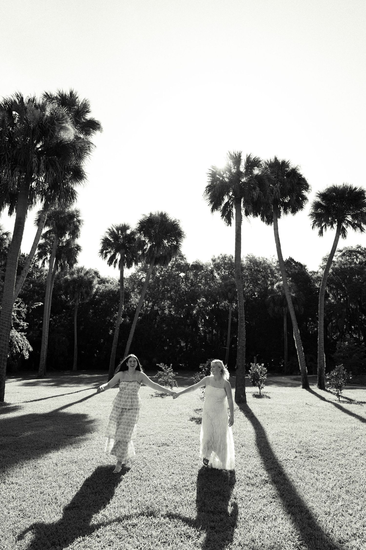 Black and white photo of two women holding hands on a lawn surrounded by tall palm trees casting long shadows.