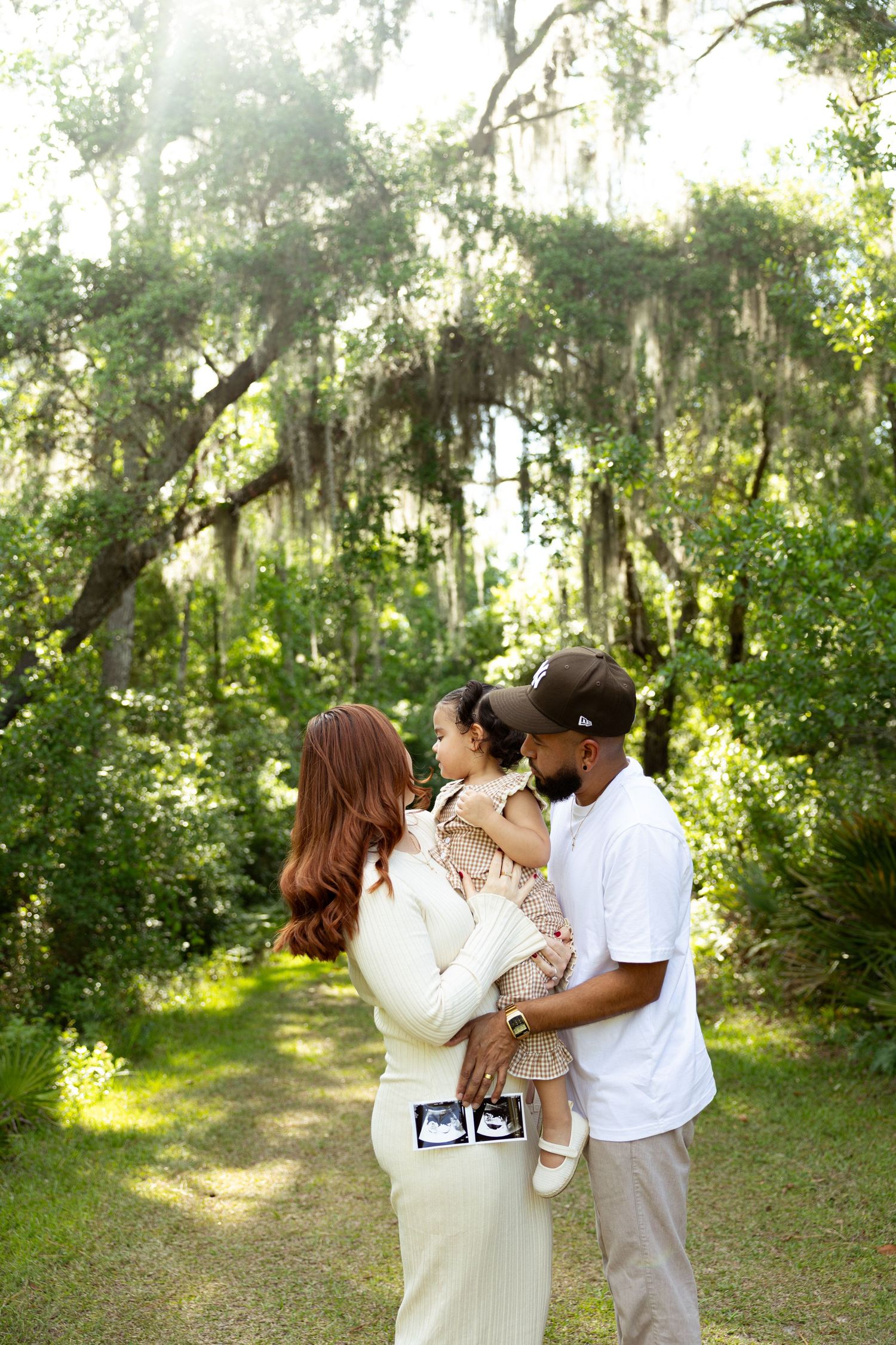 A couple holds their toddler outdoors under moss-draped trees, displaying ultrasound photos, announcing a new pregnancy.
