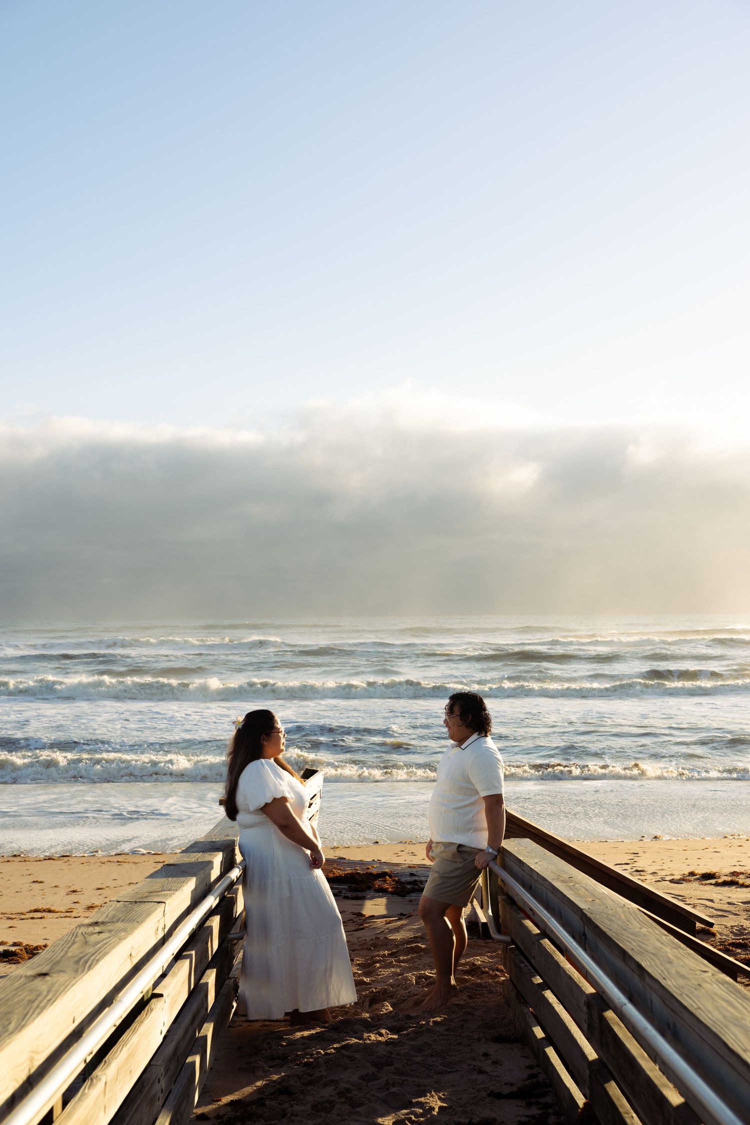 Couple in white outfits standing on a beach boardwalk, sharing a romantic moment with ocean waves in the background.