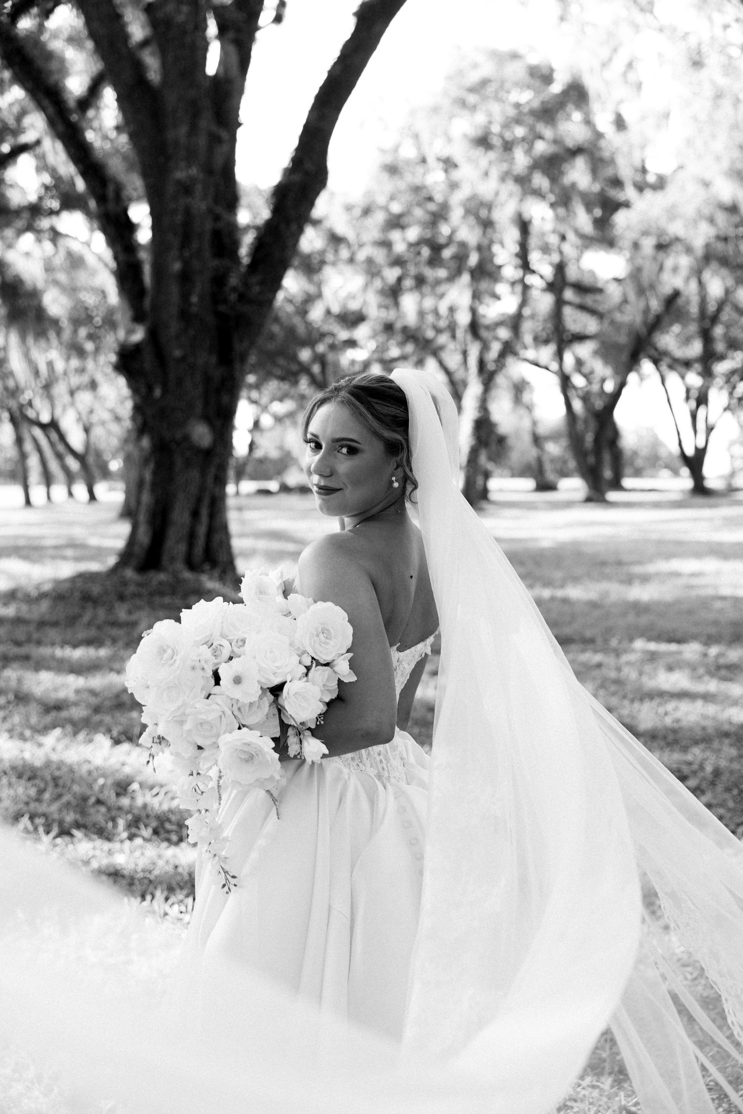 Black and white portrait of bride in strapless gown holding white bouquet with flowing veil outdoors.