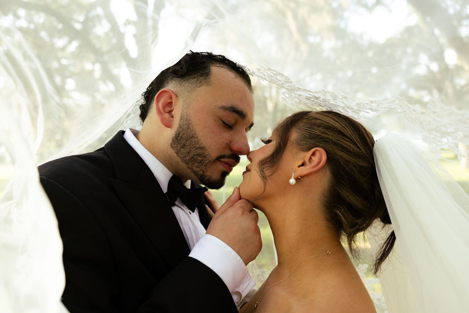 Bride and groom sharing a romantic almost-kiss under a white veil outdoors on their wedding day.
