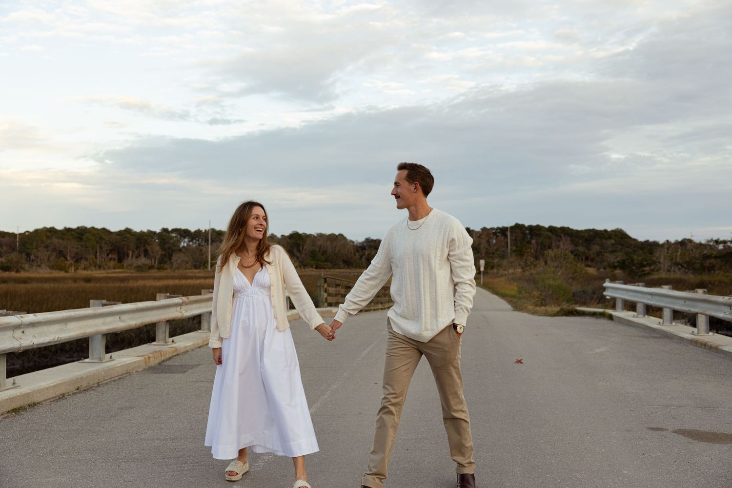 Couple holding hands on a bridge at sunset, woman in white dress, man in beige outfit, with marsh and trees behind.
