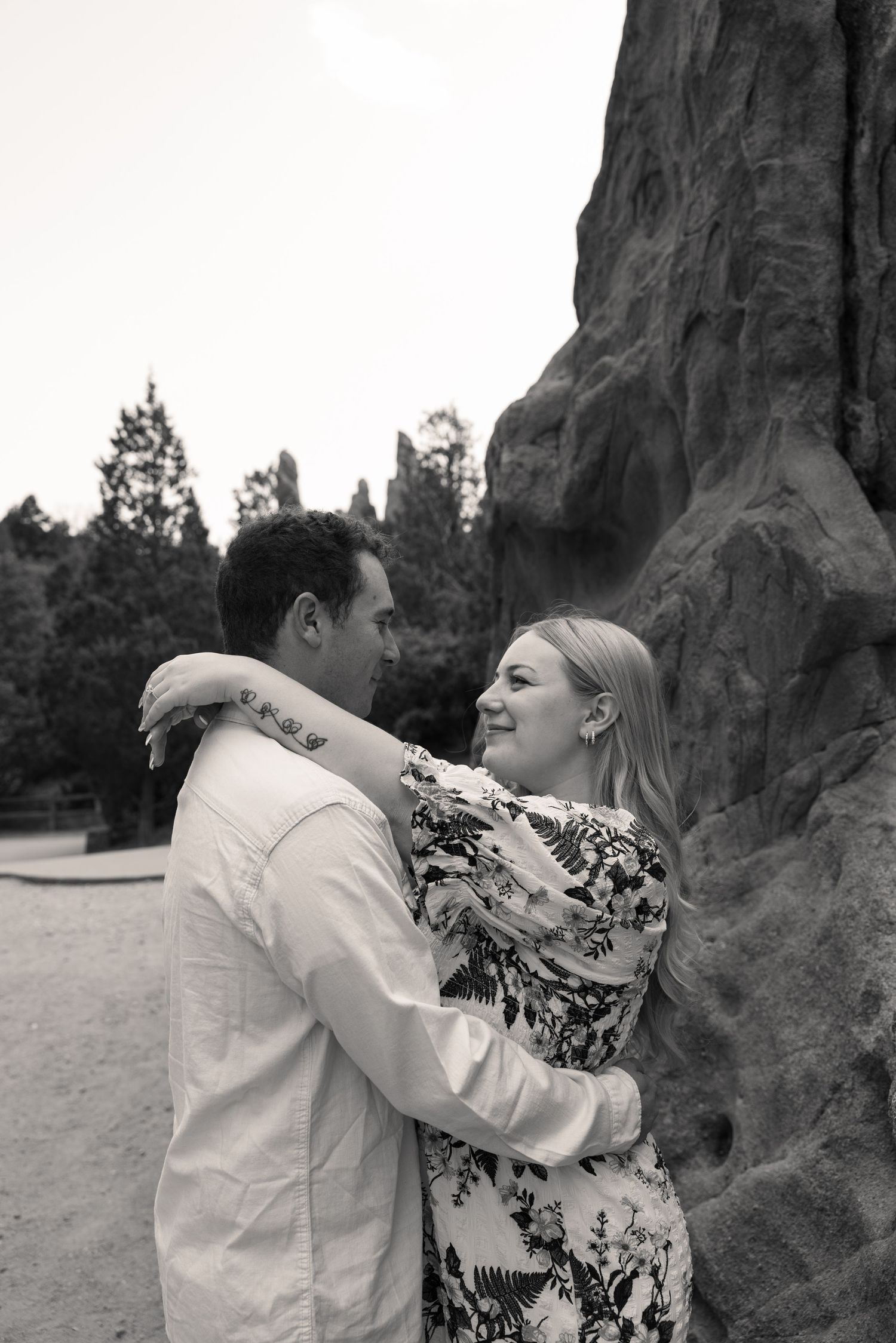 Black and white photo of a couple embracing and gazing at each other near a rocky formation outdoors.