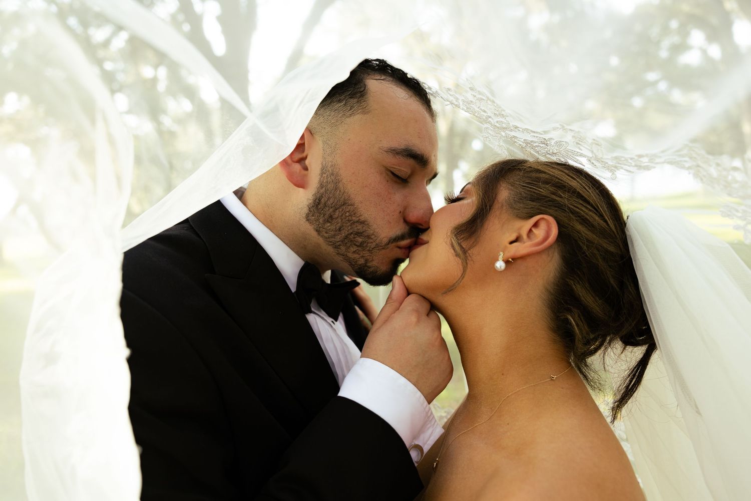 Bride and groom share a romantic kiss under a white veil on their wedding day, groom in tuxedo.