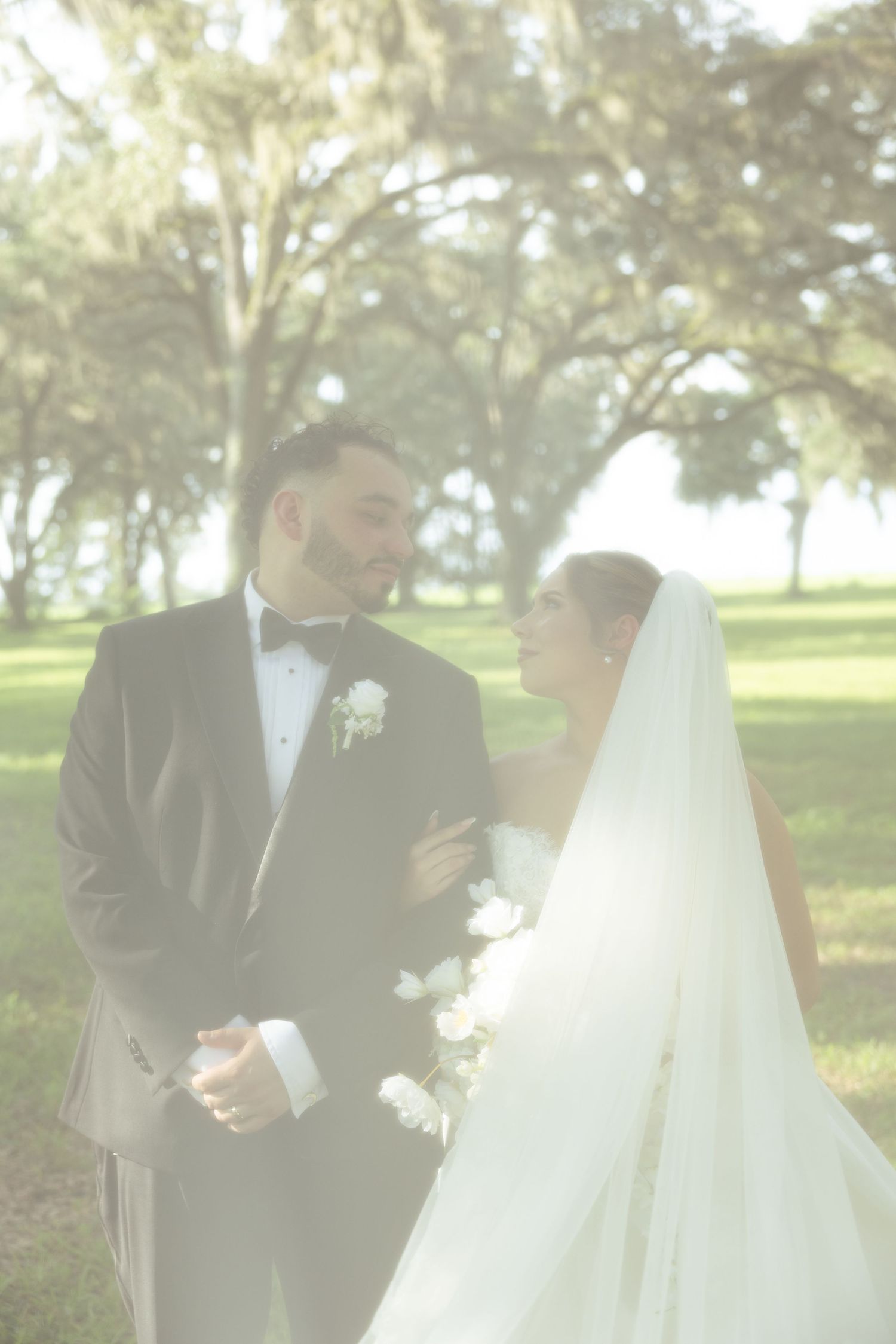 Bride and groom share an intimate moment outdoors, surrounded by lush trees in a soft, dreamy wedding portrait.