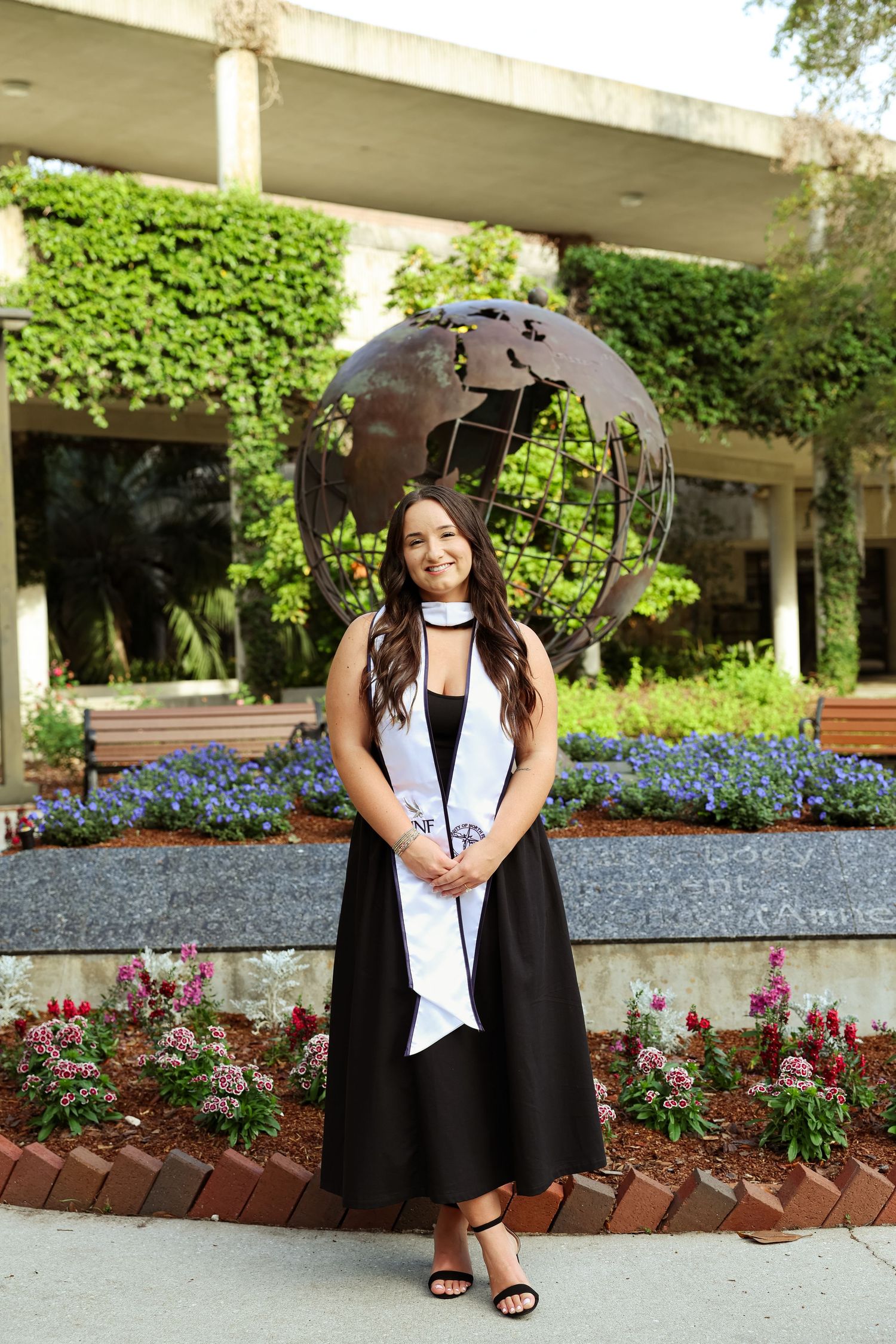 Young woman in black graduation dress with white stole poses in front of globe sculpture surrounded by colorful flowers.