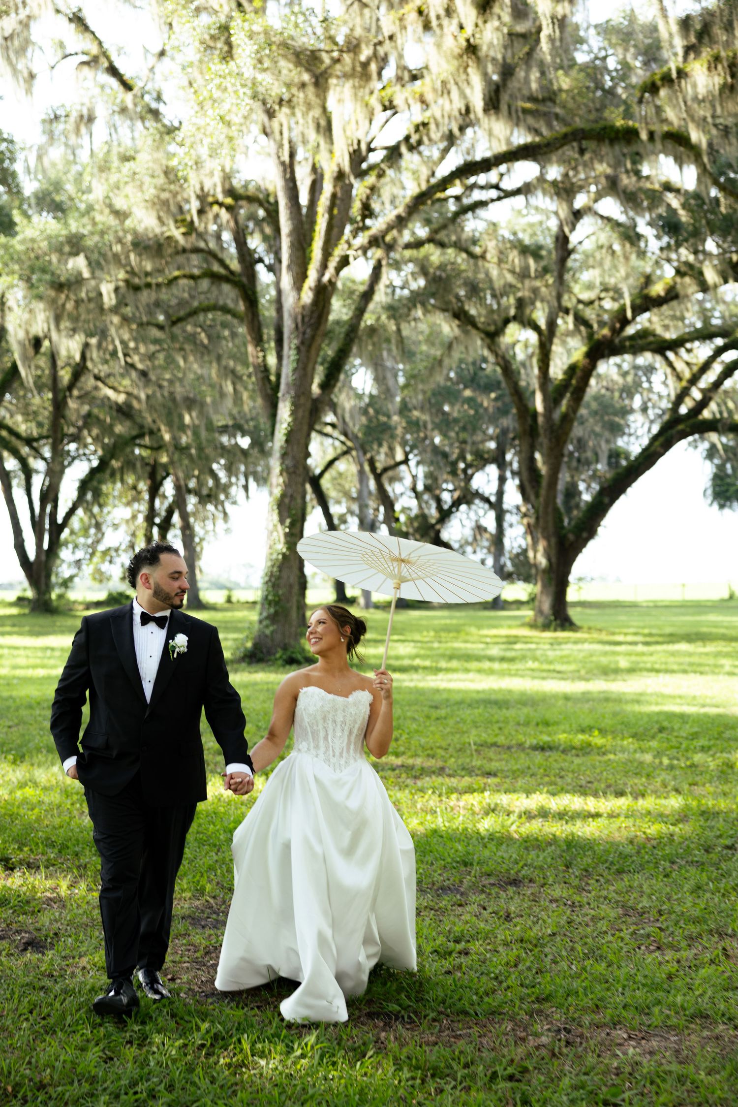 Bride and groom walk hand-in-hand under Spanish moss-draped oak trees, bride holding a white parasol in a lush green park.
