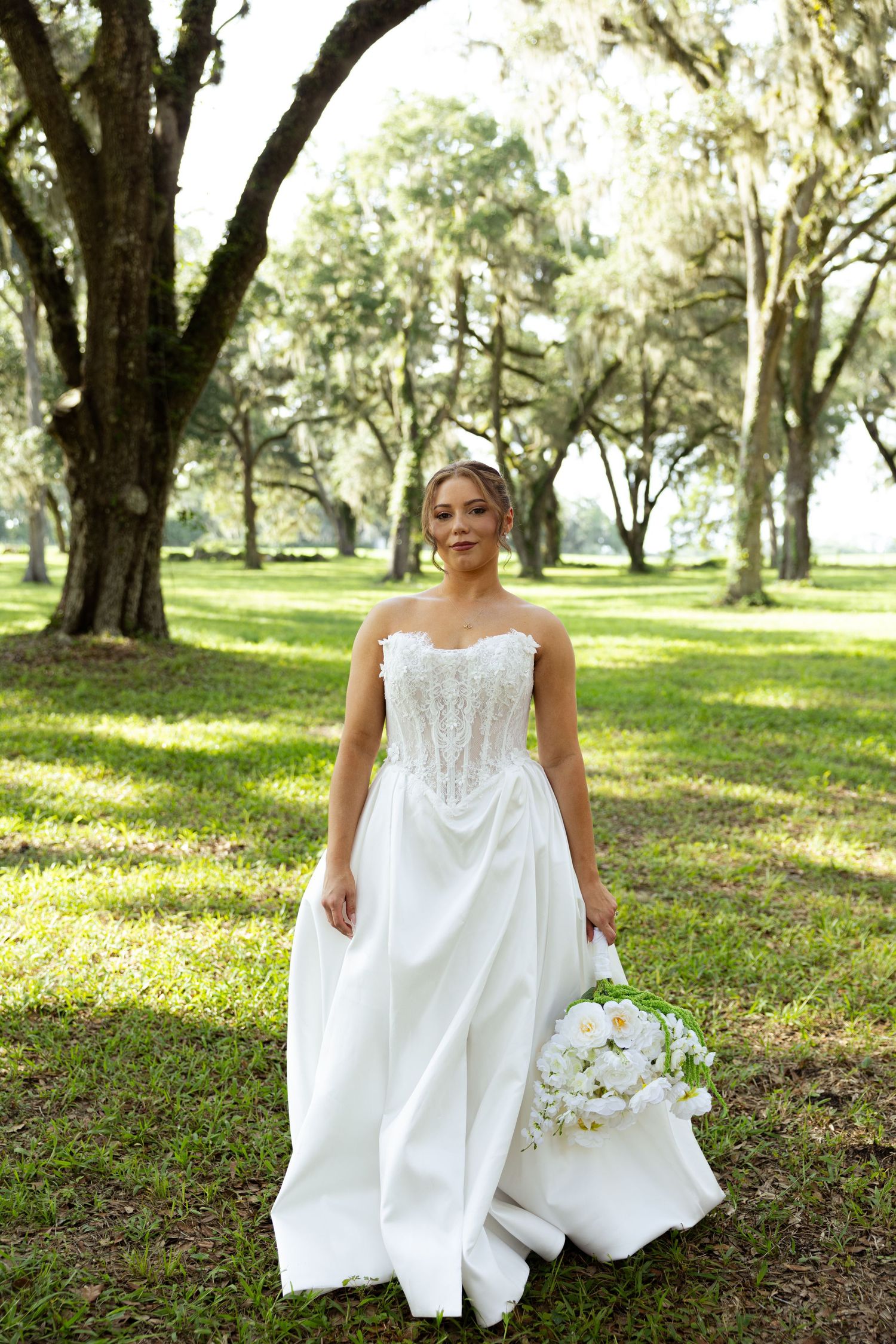 Elegant bride in strapless lace corset wedding gown holds white bouquet among moss-covered oak trees outdoors.