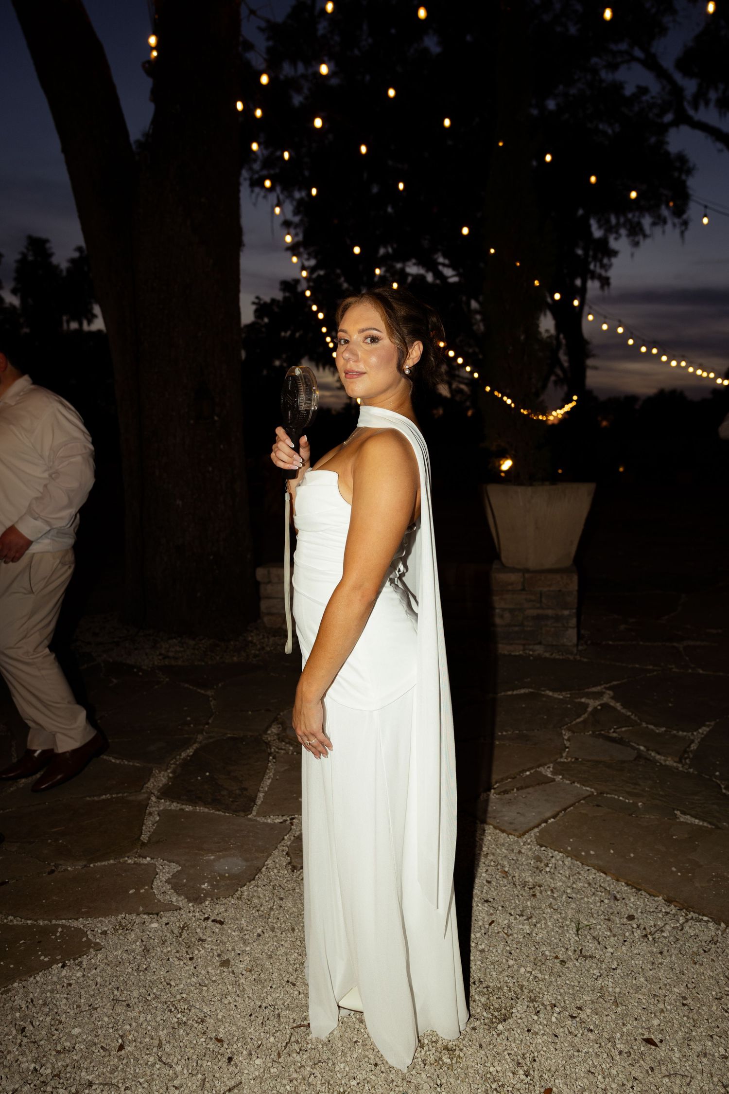 Woman in elegant white gown with black cape holds wine glass at outdoor evening wedding reception with string lights.