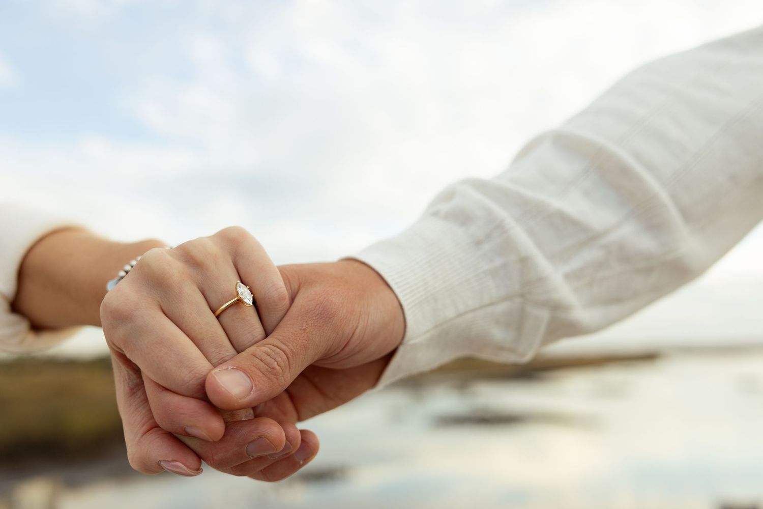 Close-up of couple holding hands with elegant gold wedding ring, romantic beach background.