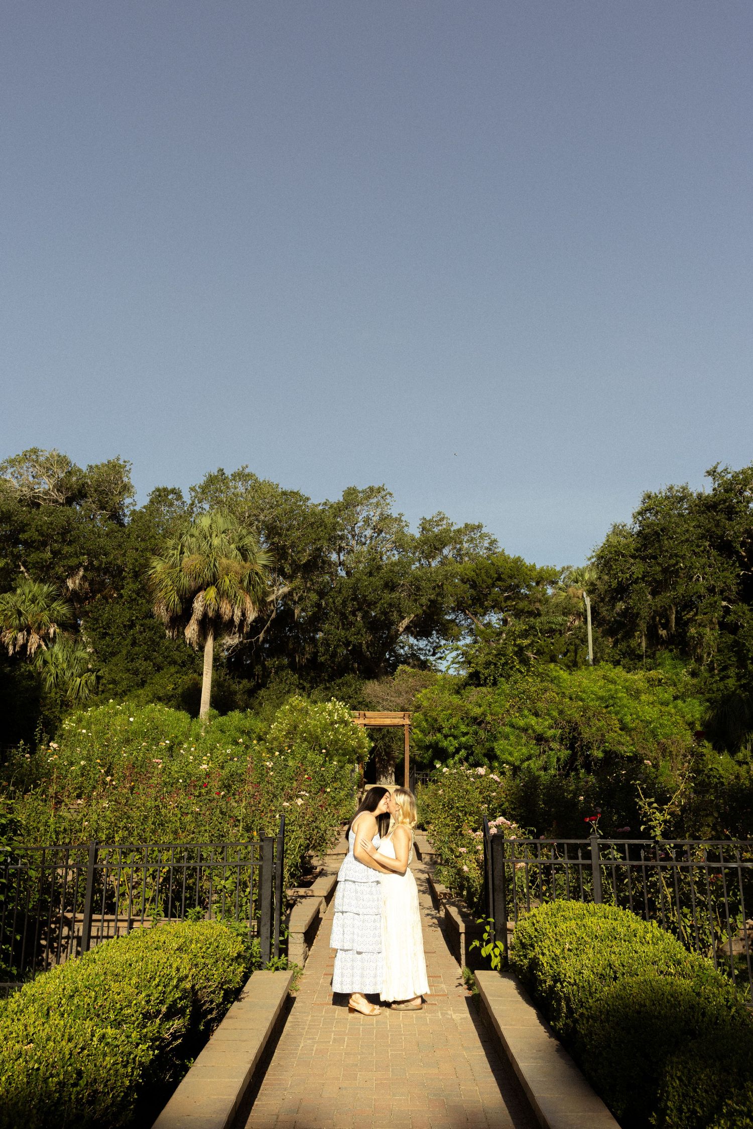 Couple in white attire embracing on a garden path surrounded by lush greenery and a gazebo under a clear blue sky.