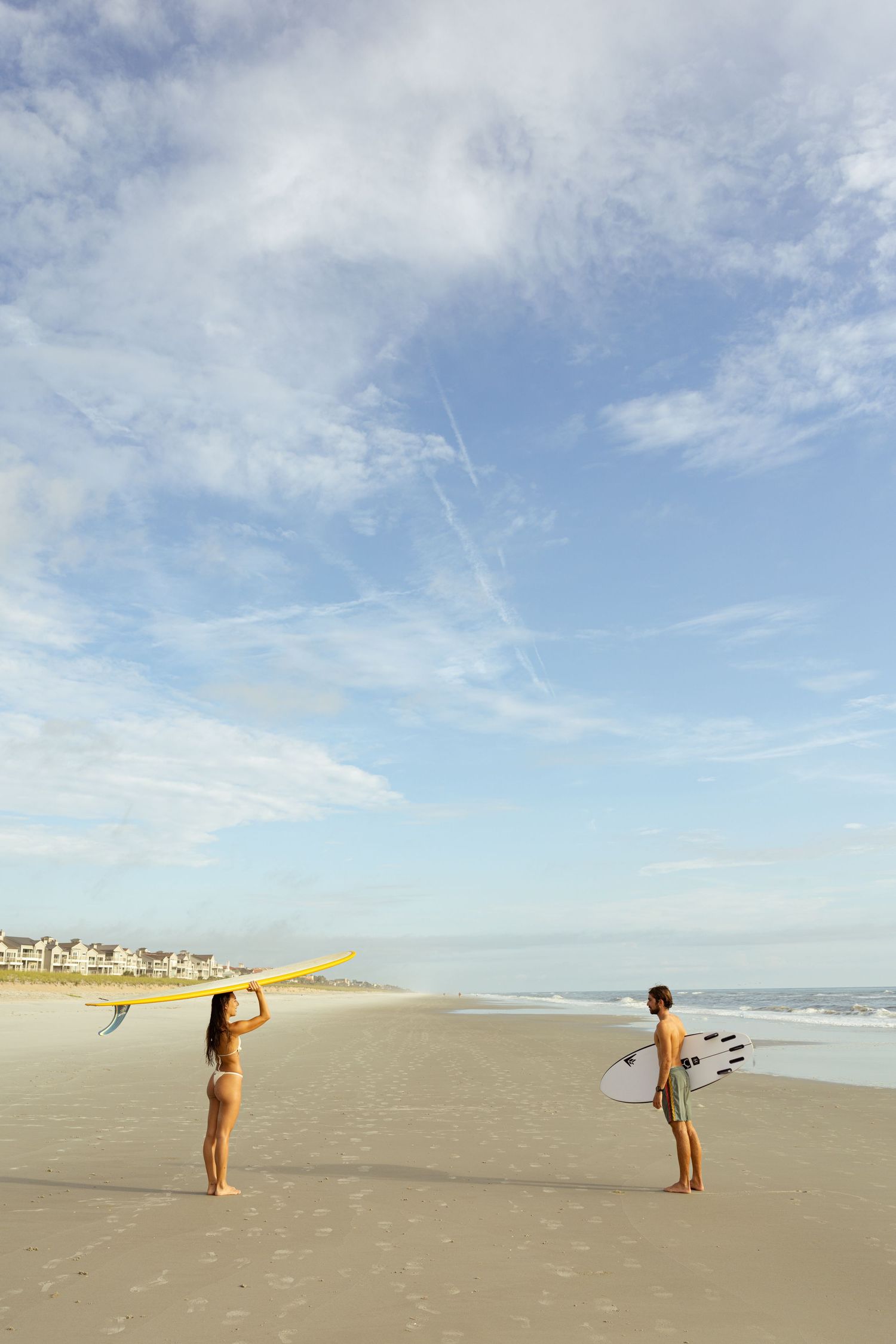 Two surfers holding boards on a sandy beach with ocean waves and cloudy blue sky in the background.