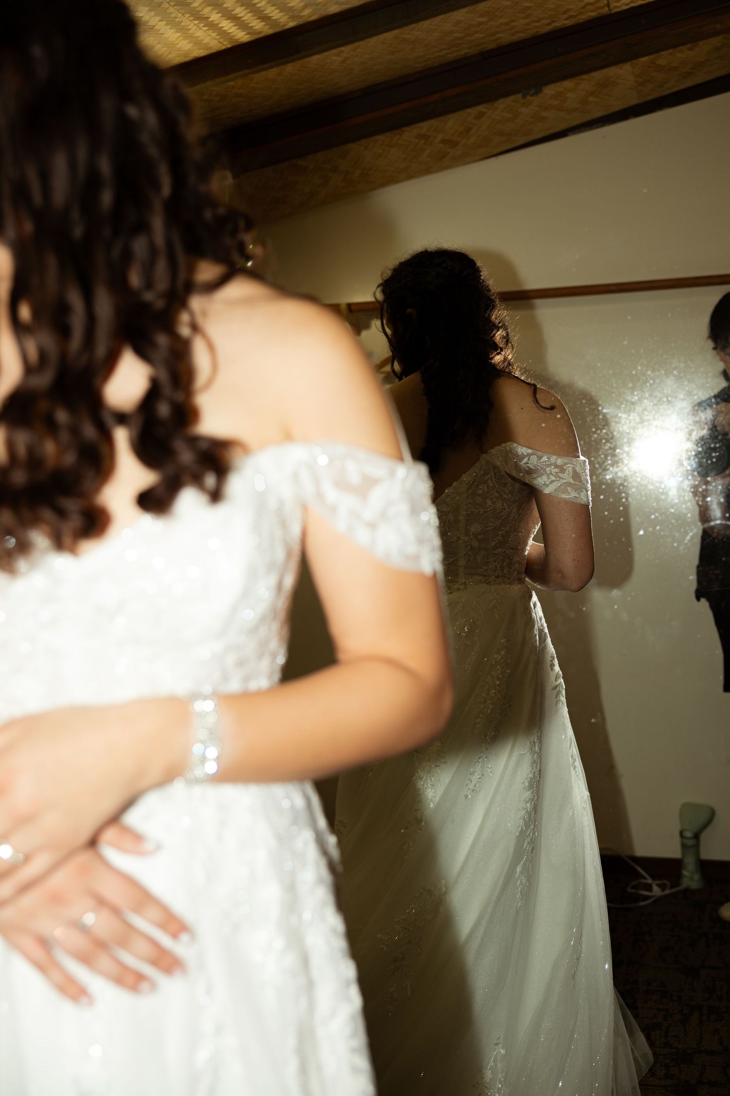 Bride in white off-shoulder lace gown views her reflection in mirror with flash photography at wedding venue.