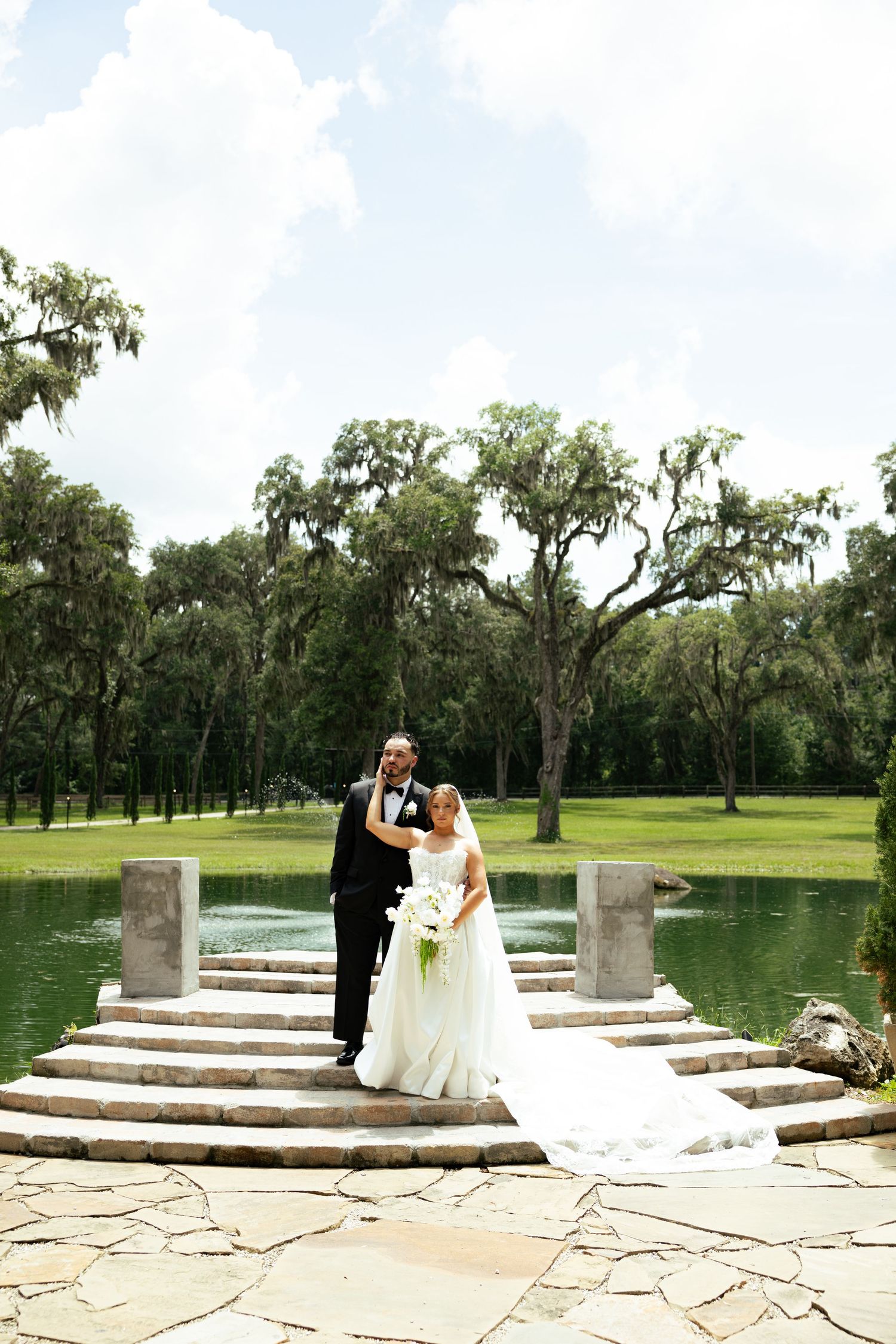 Bride and groom pose on stone steps by a serene pond surrounded by lush trees on their wedding day.
