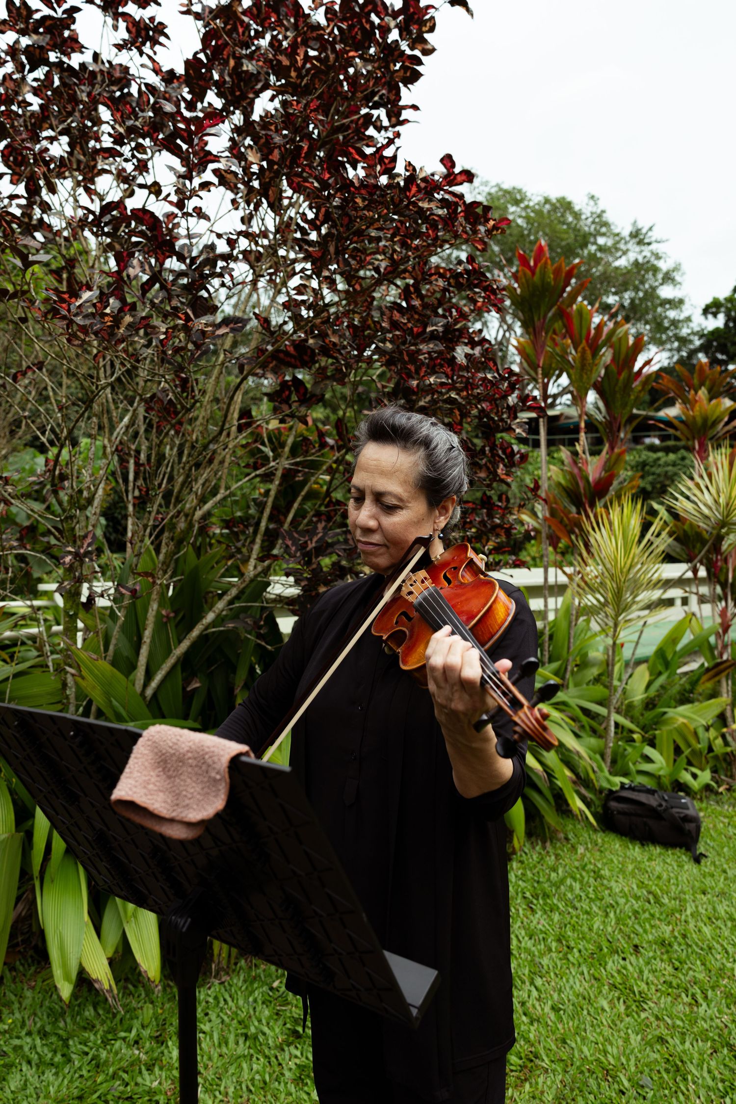 Woman in black outfit playing violin outdoors in a lush garden with a music stand nearby.