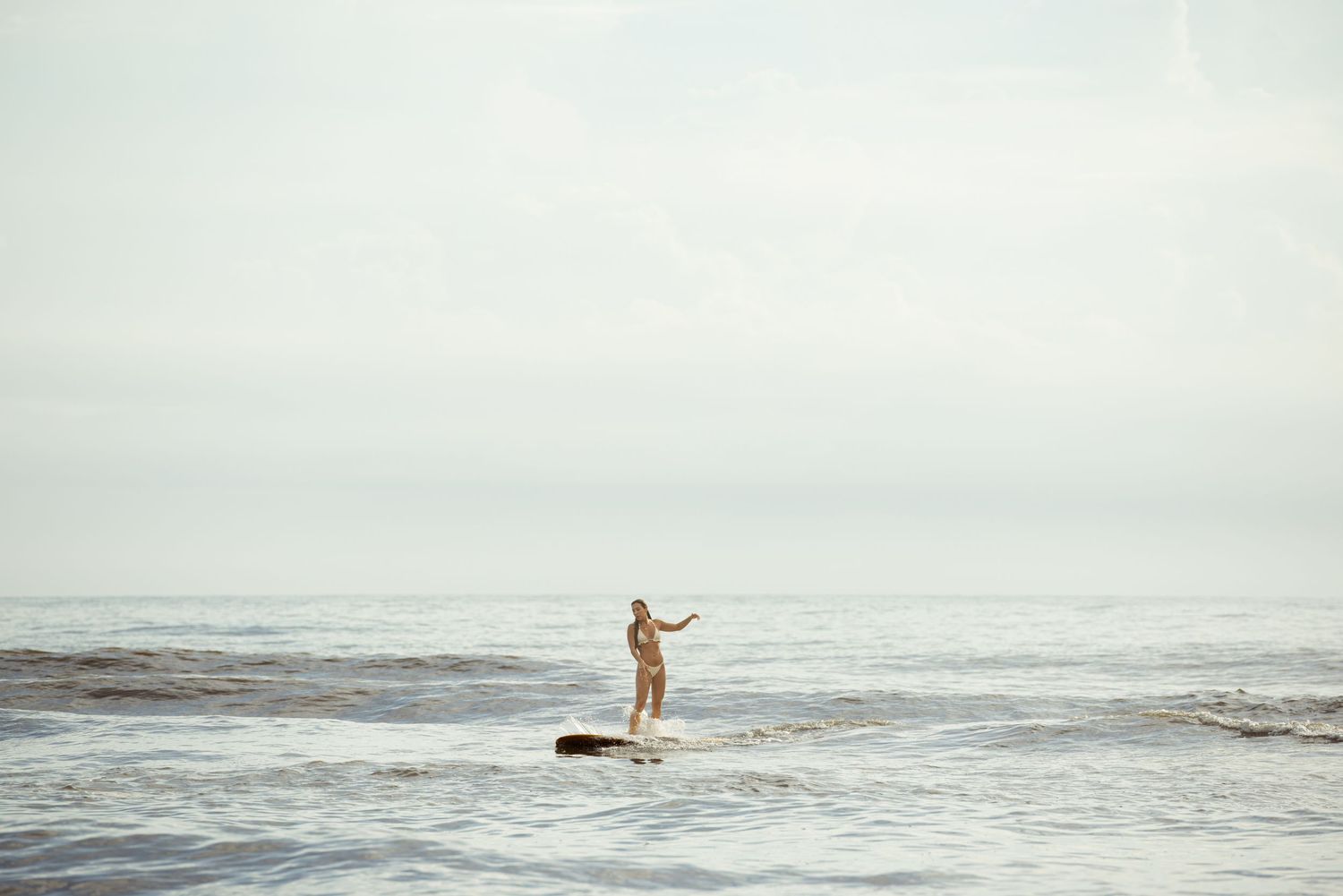 A surfer stands balancing on a surfboard in calm ocean waves under a pale overcast sky.