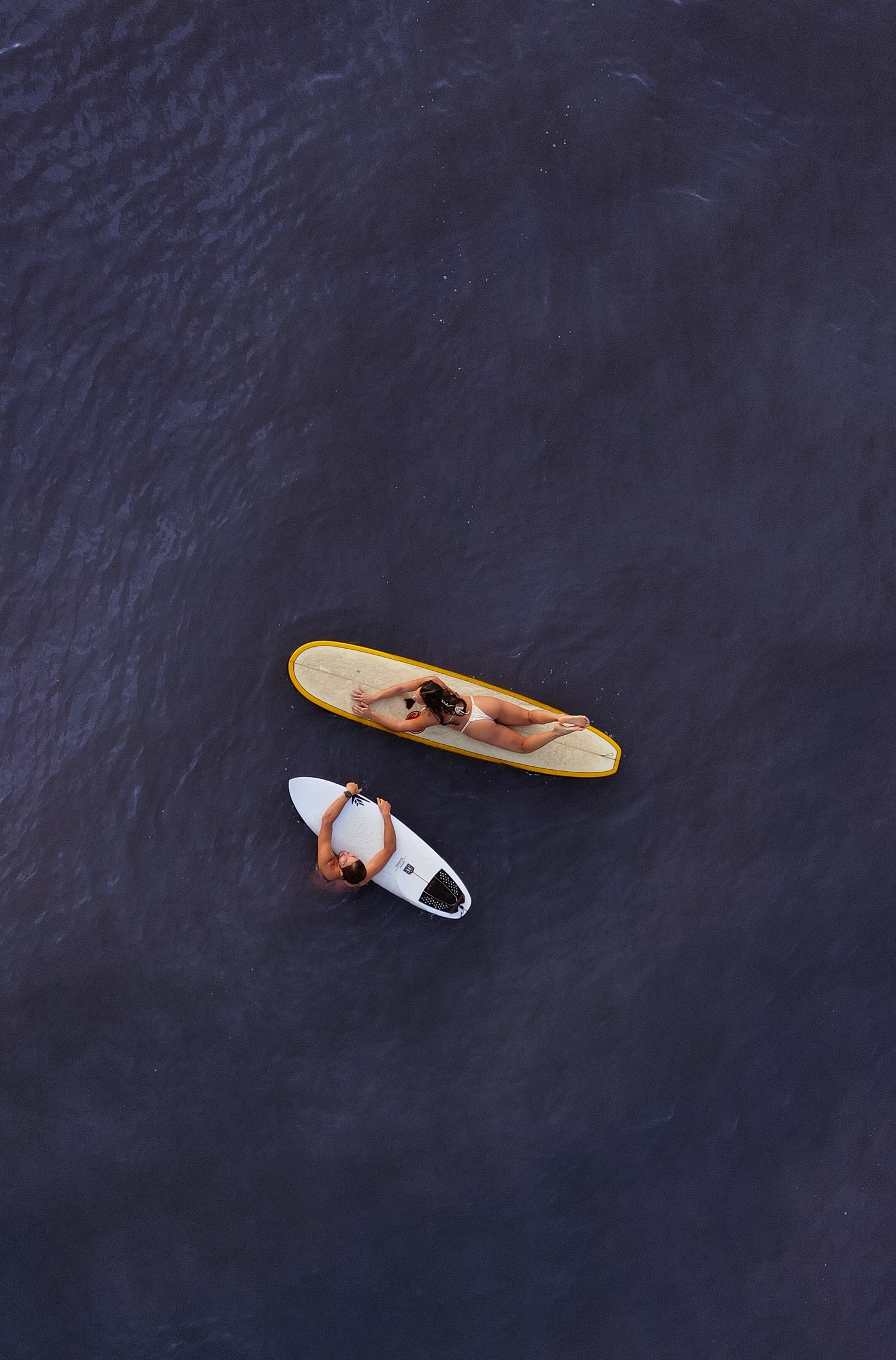 Aerial view of two surfers lying on surfboards floating on calm dark ocean water.