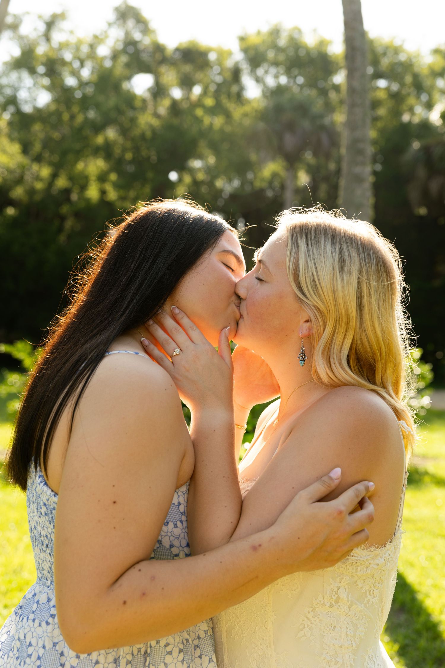 Two women sharing a romantic kiss outdoors in a sunlit garden setting.
