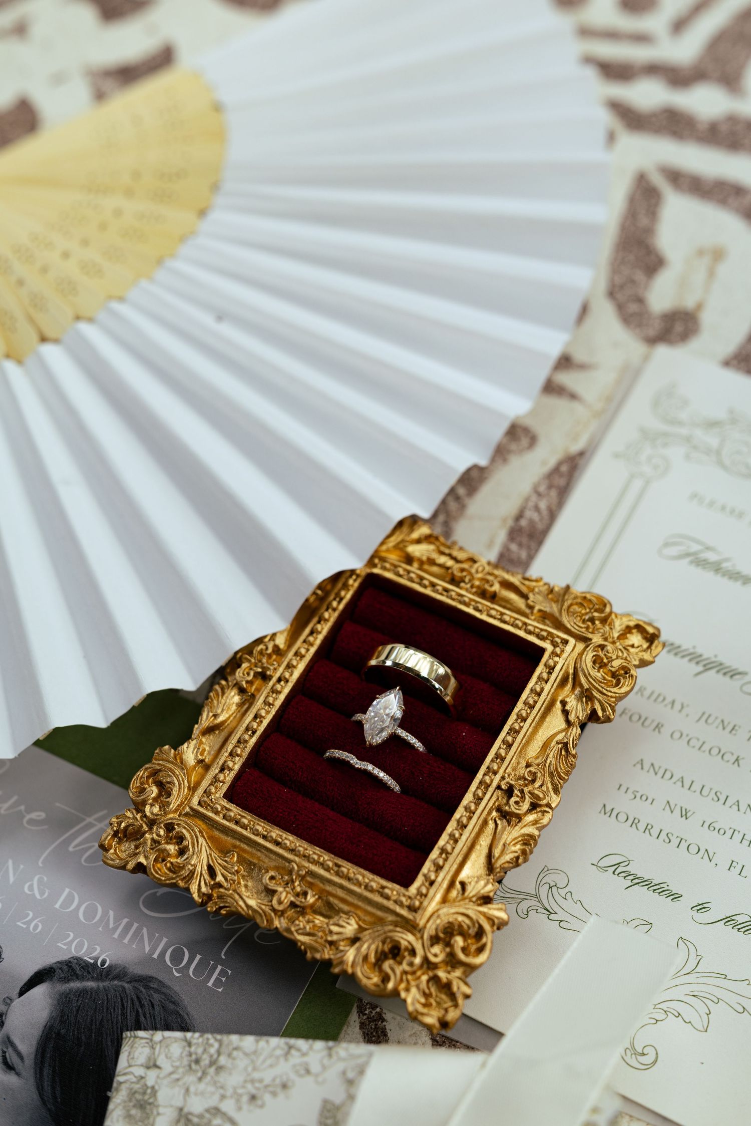 Gold ornate ring box with diamond engagement and wedding bands displayed with fan and wedding invitations.