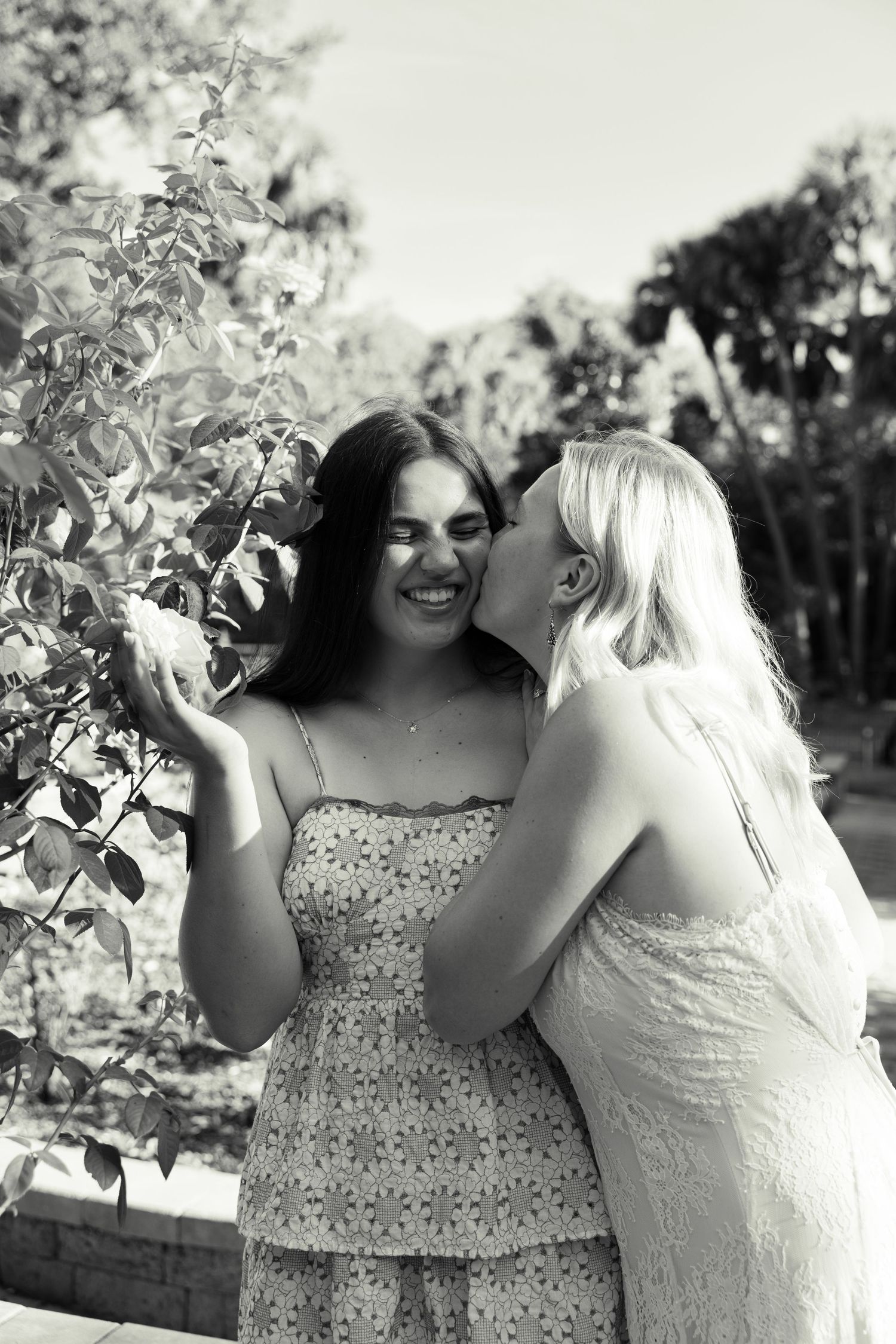 Black and white photo of two young women outdoors, one kissing the other on the cheek near leafy green plants.