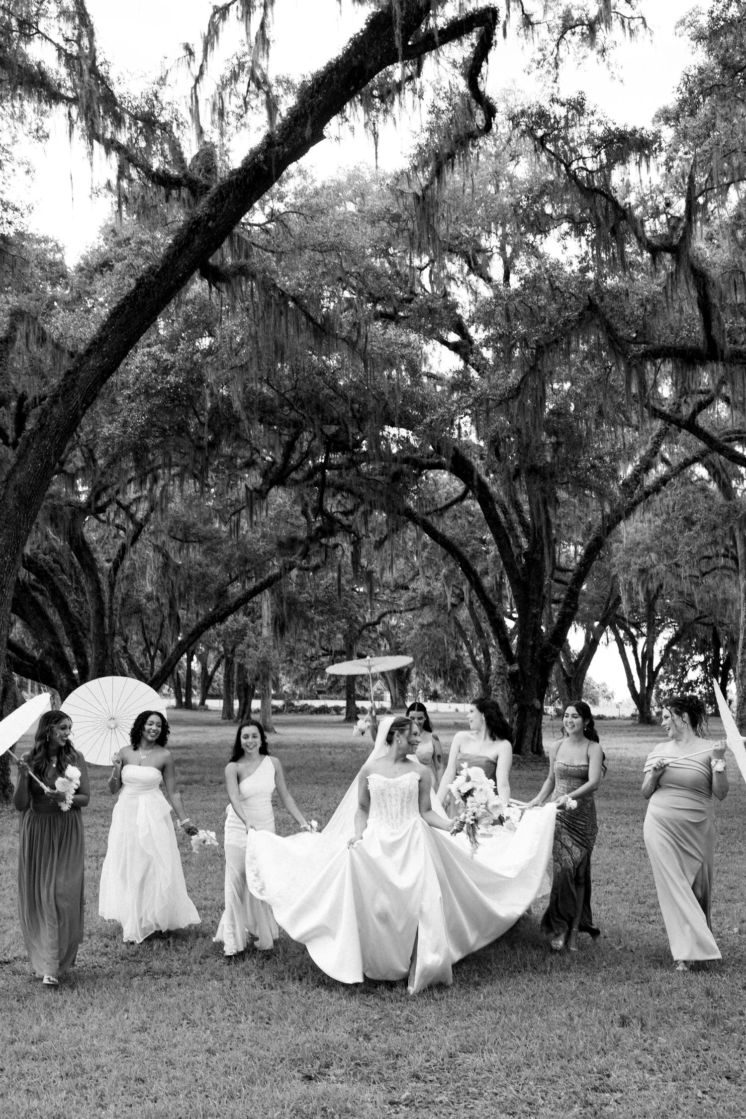 Black and white photo of a bride and bridesmaids walking outdoors under moss-draped oak trees at a wedding.