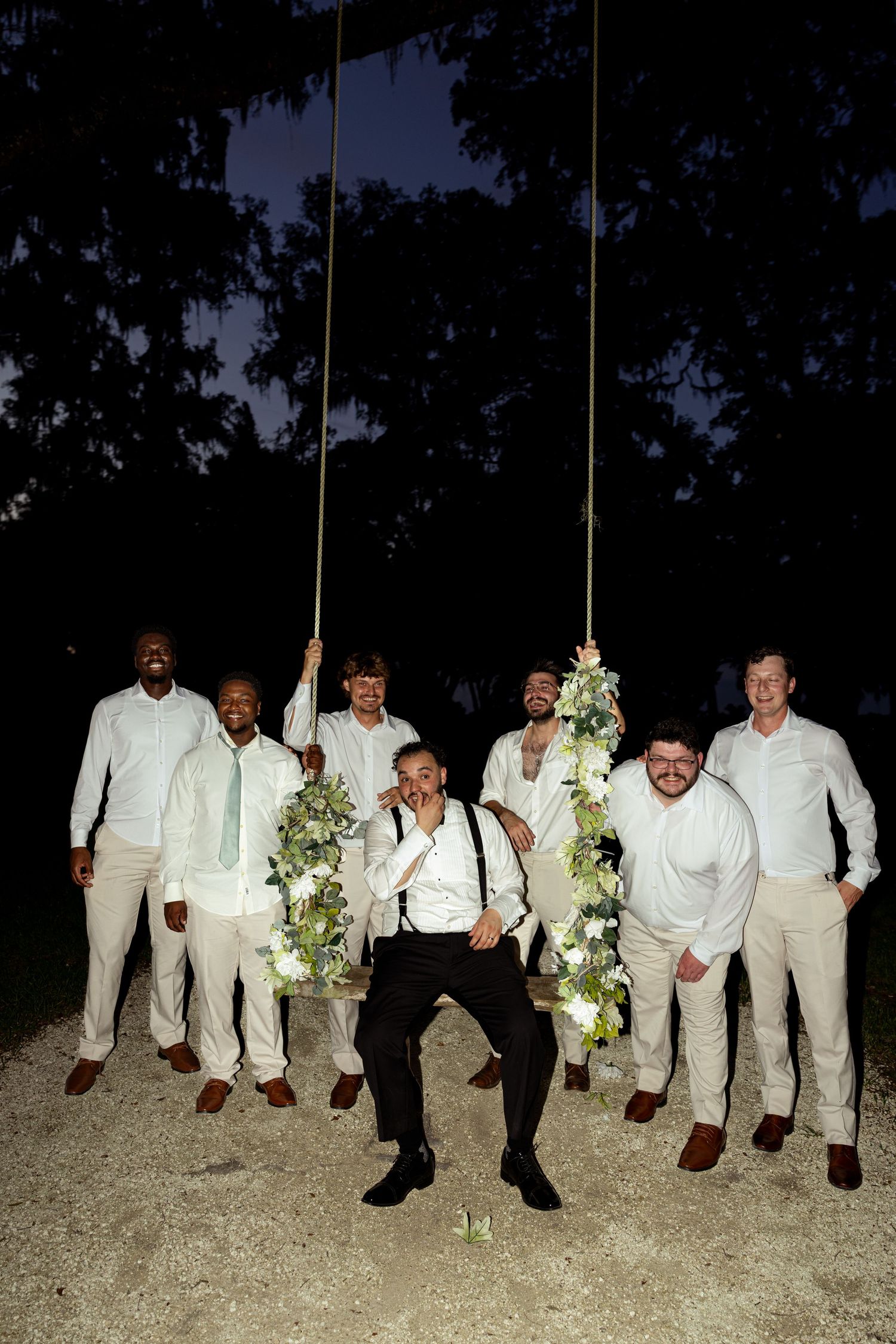 Groom sits on floral swing surrounded by groomsmen in white shirts and khakis during outdoor evening wedding celebration.
