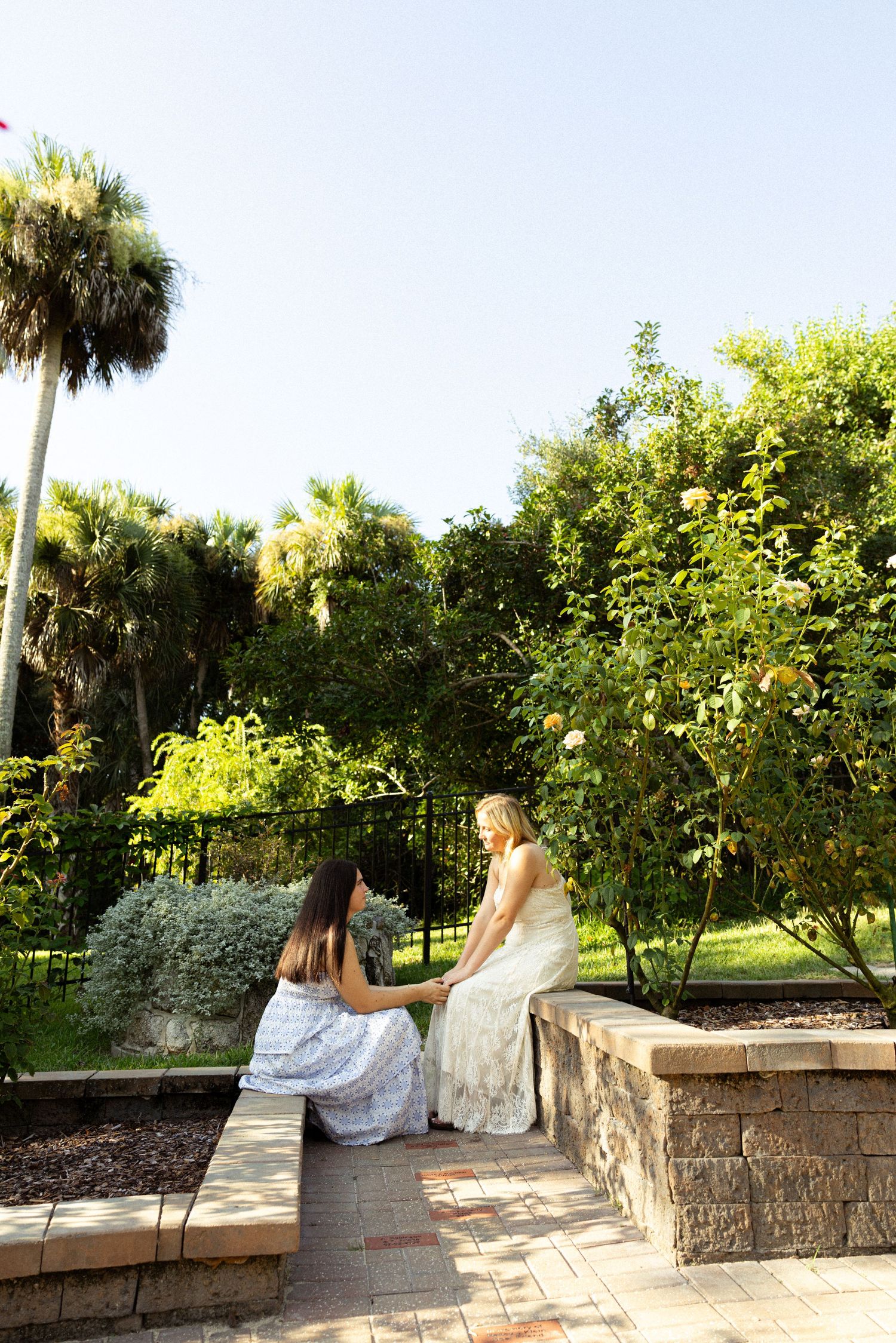 Two women in white dresses sitting together on a stone wall in a lush garden setting.