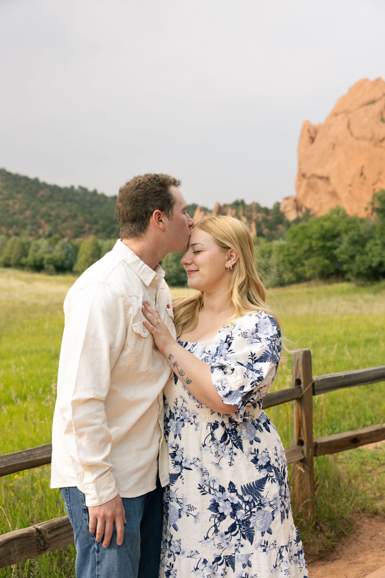 Couple sharing a tender kiss near a wooden fence with red rock formations and green meadow in the background.